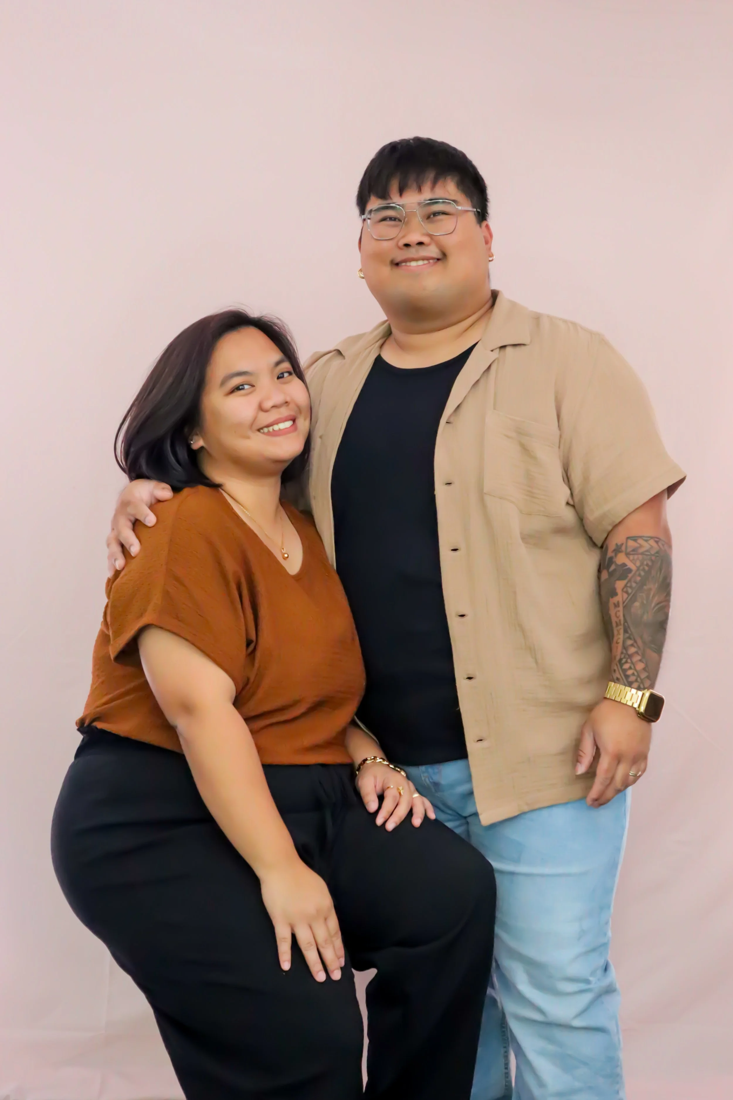 A smiling man and woman posing together against a plain light pink background, with the woman sitting and the man standing. They are close, with the man's arm around the woman's shoulder.