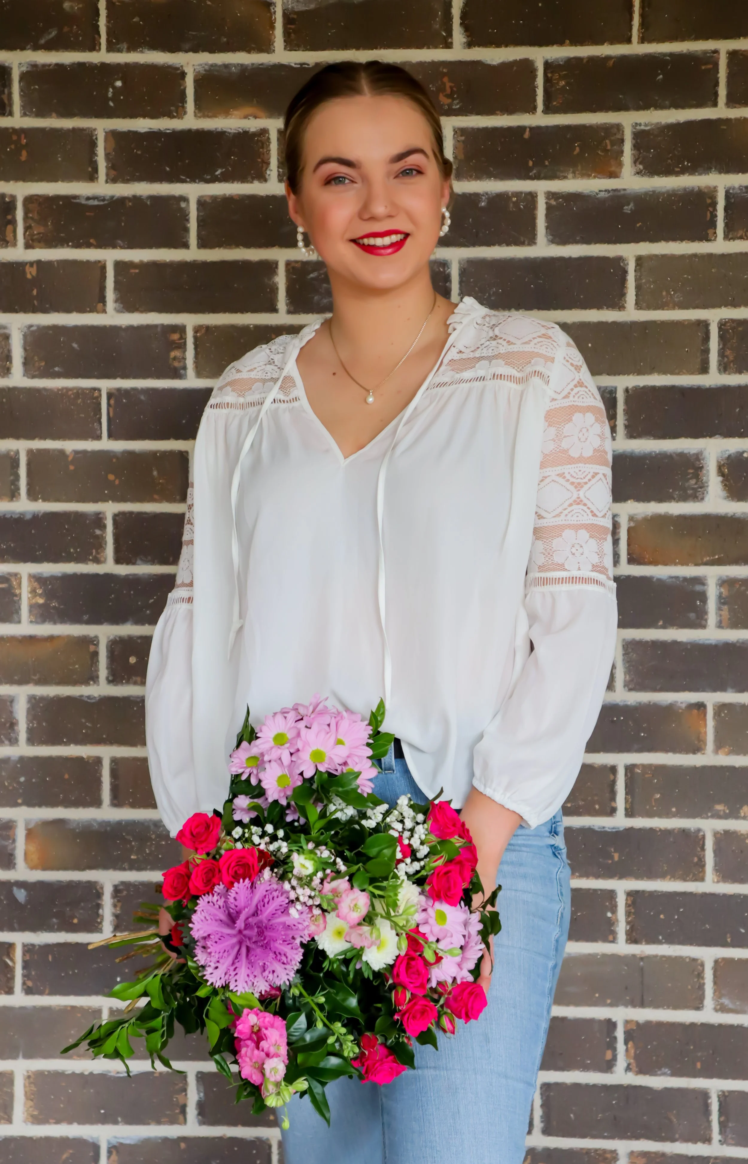 A young woman with brown hair wearing a white blouse and blue jeans holding a large bouquet of pink, purple, and red flowers, standing in front of a brick wall.