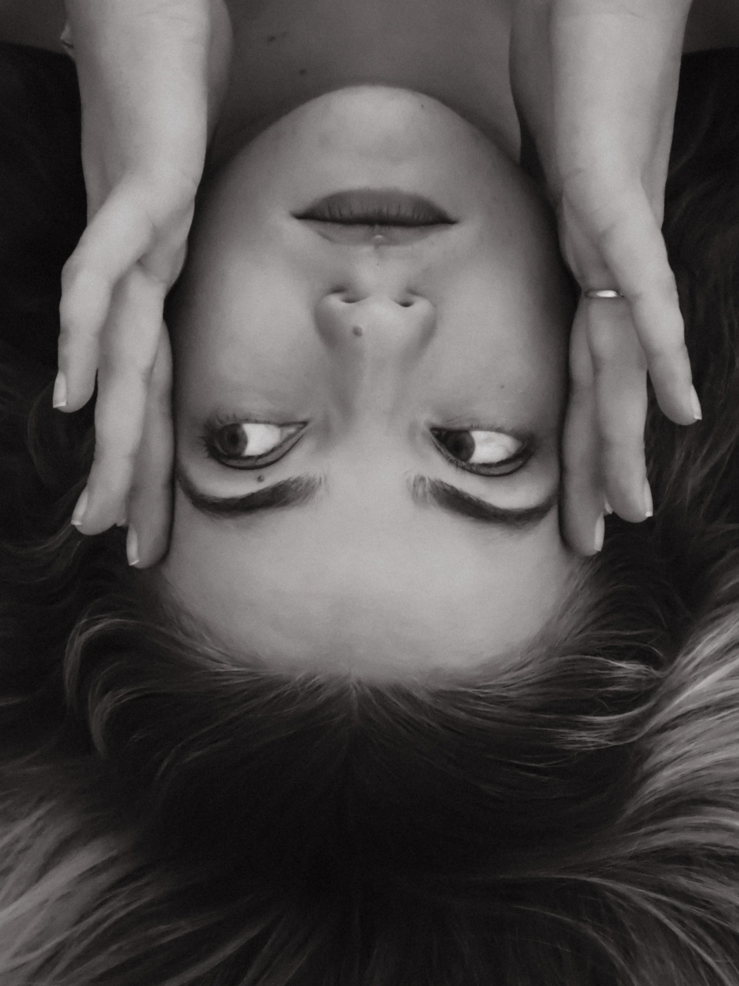 A woman lying down with her head upside down, features her hands framing her face, staring towards the camera with a calm expression, in black and white.