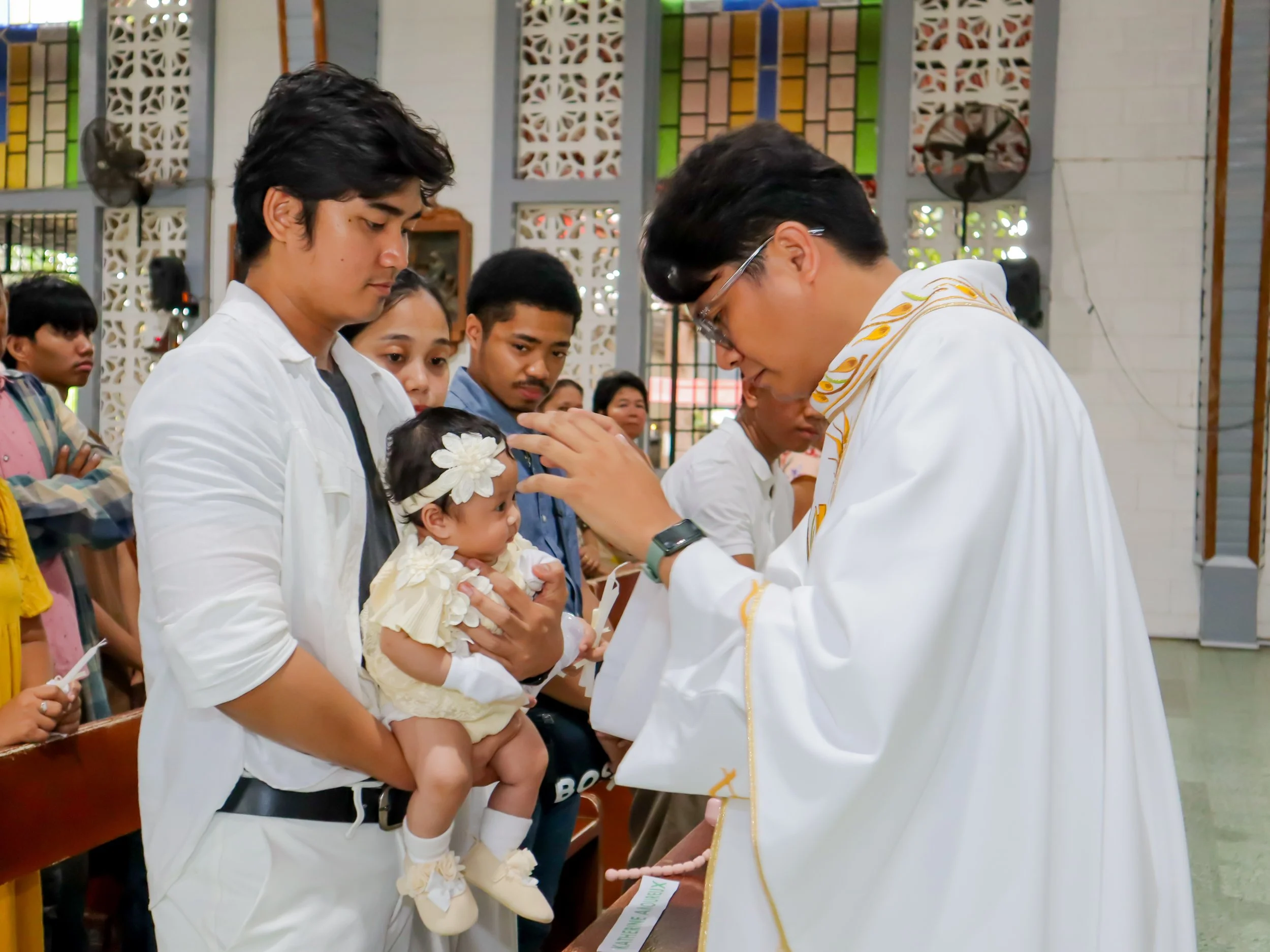 A priest administering a religious blessing or sacrament to a baby held by a young man during a church ceremony, with several people observing in the background.
