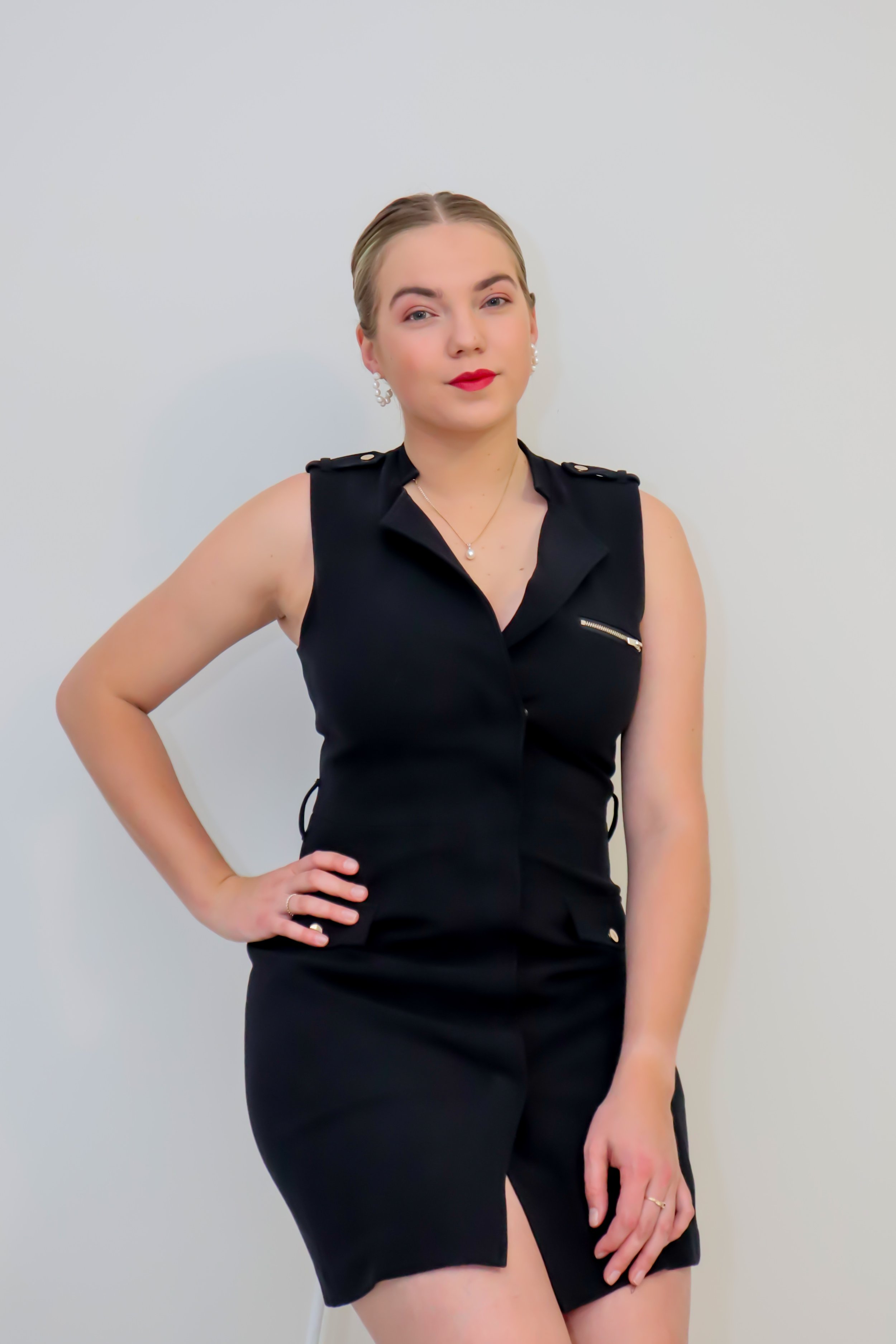 Woman wearing a black sleeveless dress with gold buttons and zipper, standing with one hand on her hip and the other resting on her thigh, against a plain white wall.