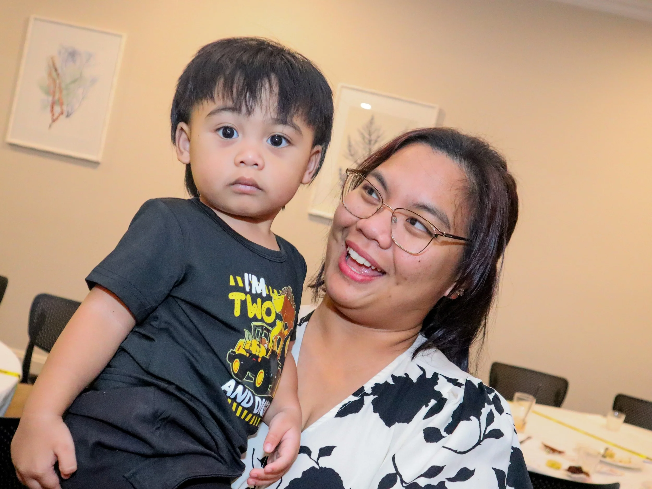 A woman with glasses holding a young boy with dark hair and big eyes, both smiling in an indoor setting with artwork on the wall behind them.