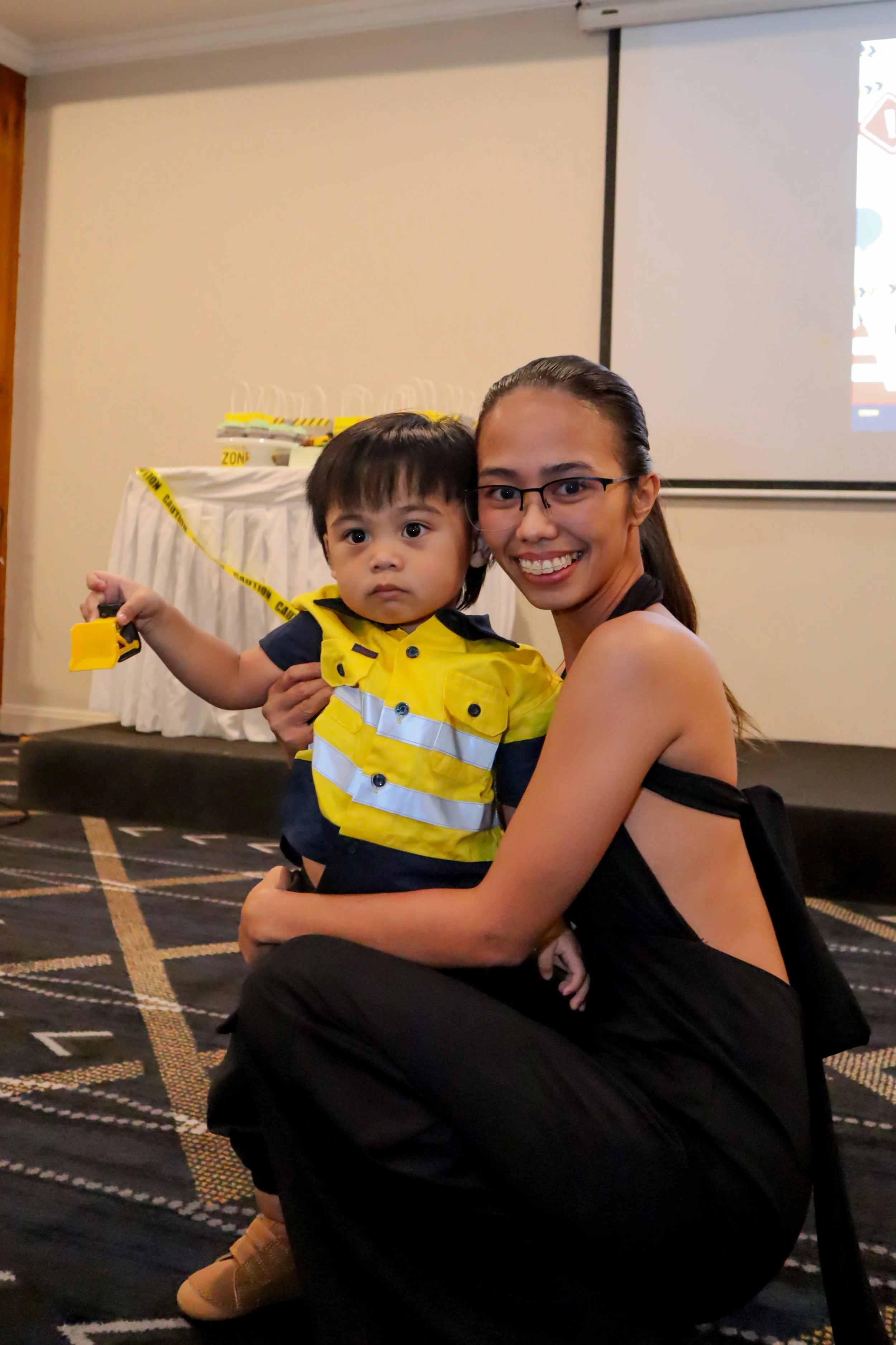 A woman and a young boy sitting on the floor. The boy is wearing a yellow safety vest and holding a yellow toy. The woman is smiling and wearing glasses and a black dress. In the background, there is a table with yellow caution tape and safety equipm