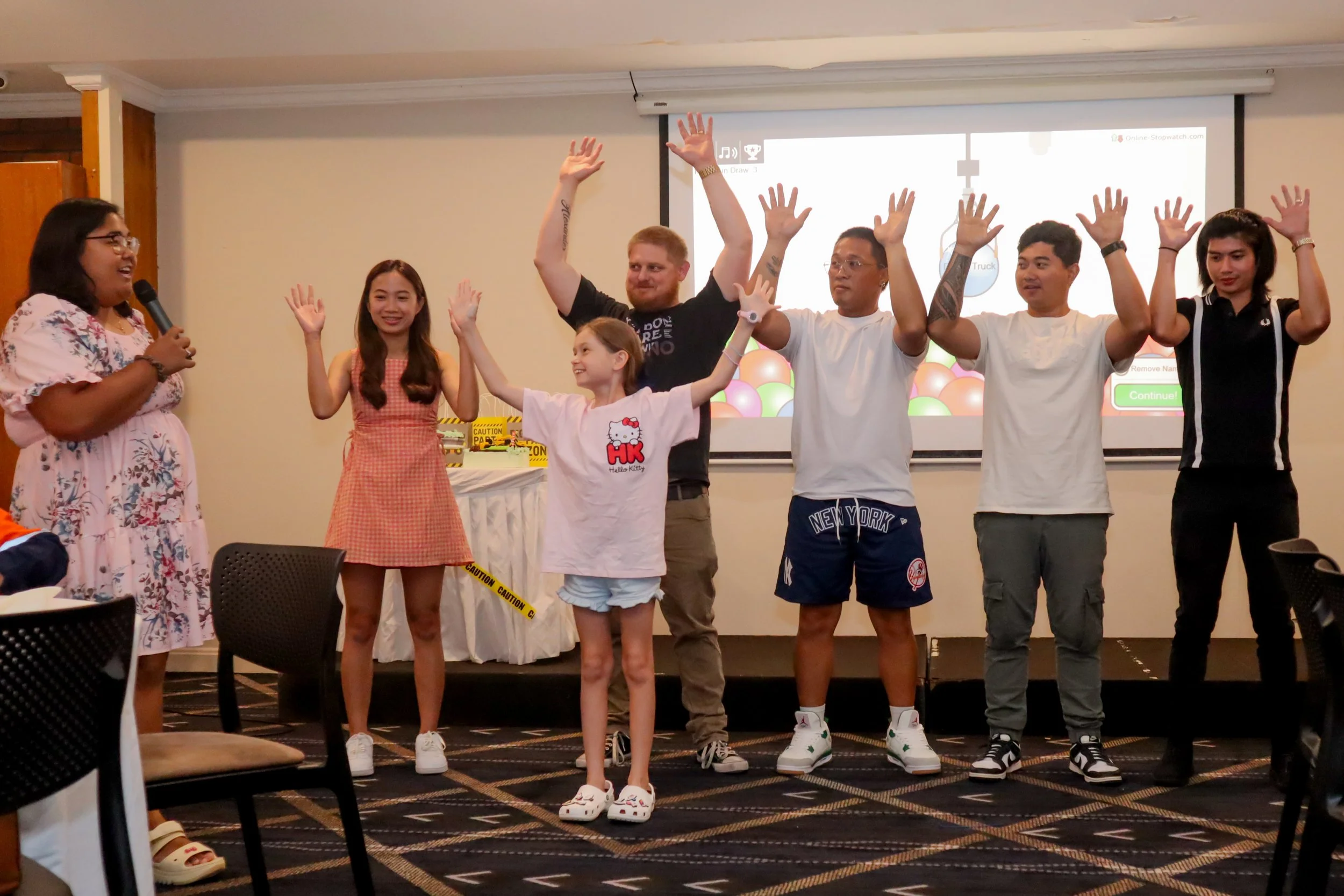 A group of seven children and teenagers standing on a stage, with one woman speaking into a microphone. The group has their hands raised in the air, and they are smiling. Behind them is a large screen displaying colorful graphics and caution tape on 