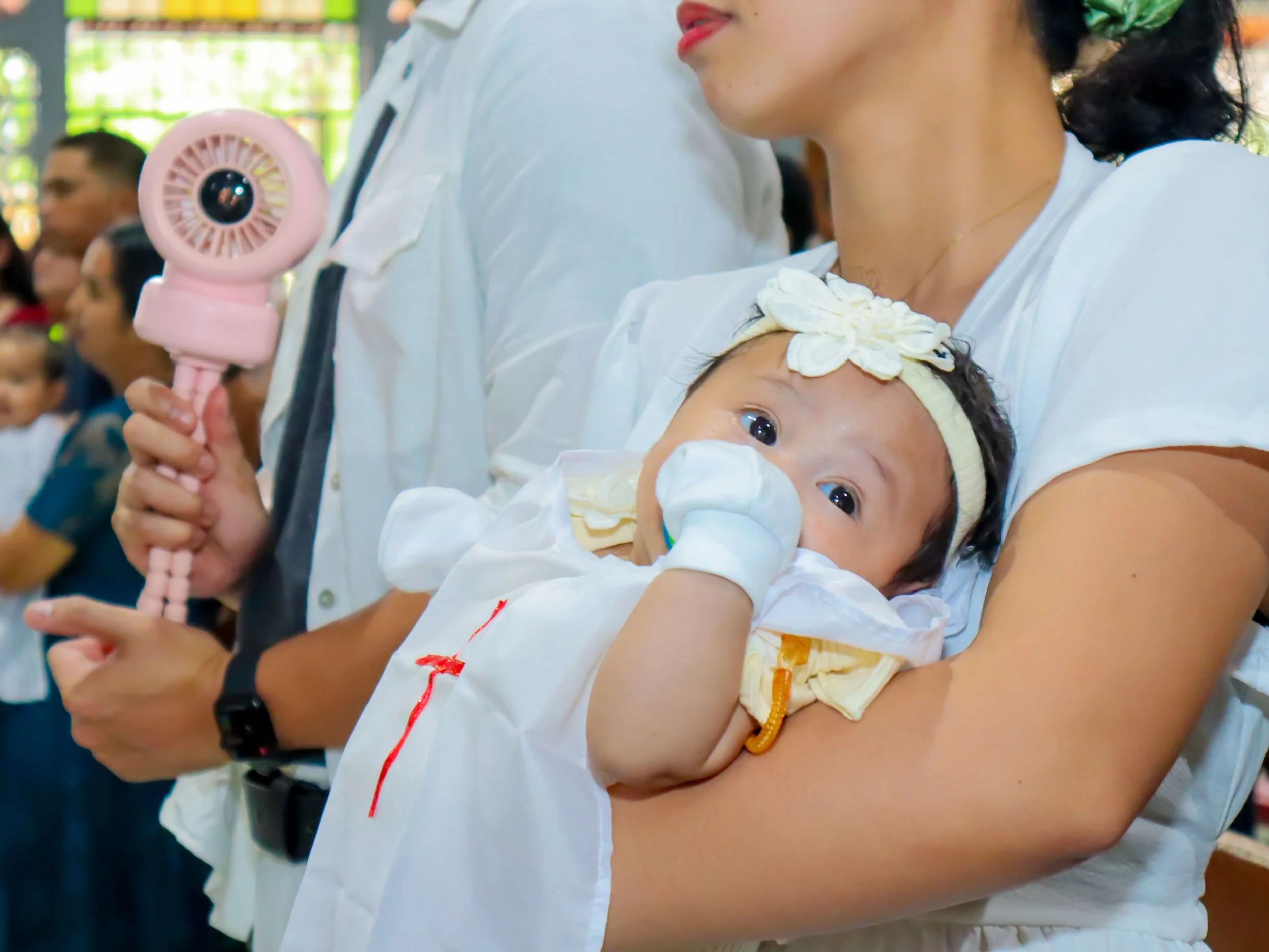 A woman holding a baby dressed in white during a public event, with a man beside her holding a small pink fan. Other people are visible in the background.