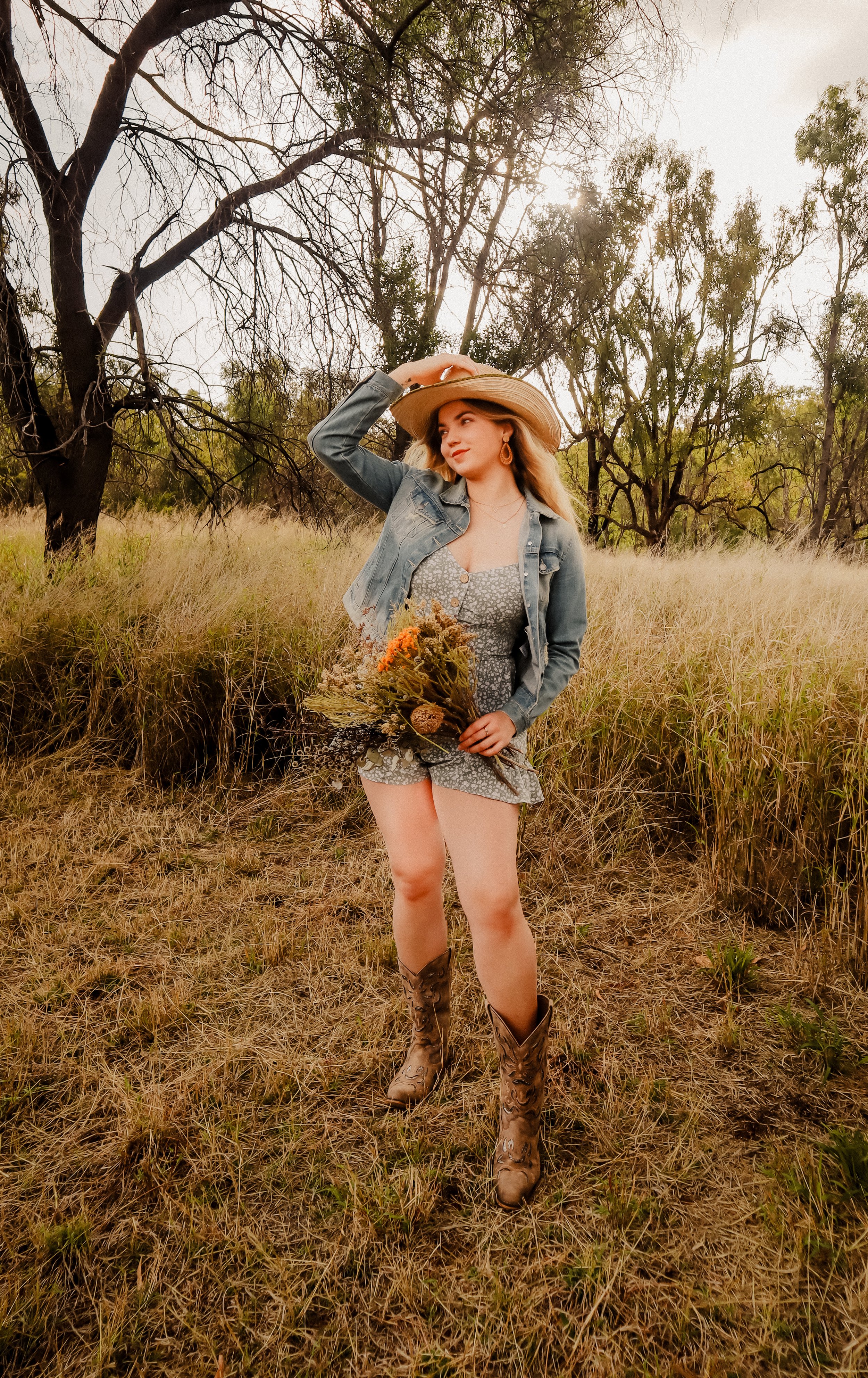 A woman stands in a grassy field with trees in the background, wearing a cowboy hat, denim jacket, floral romper, and cowboy boots, holding a bouquet of wildflowers.