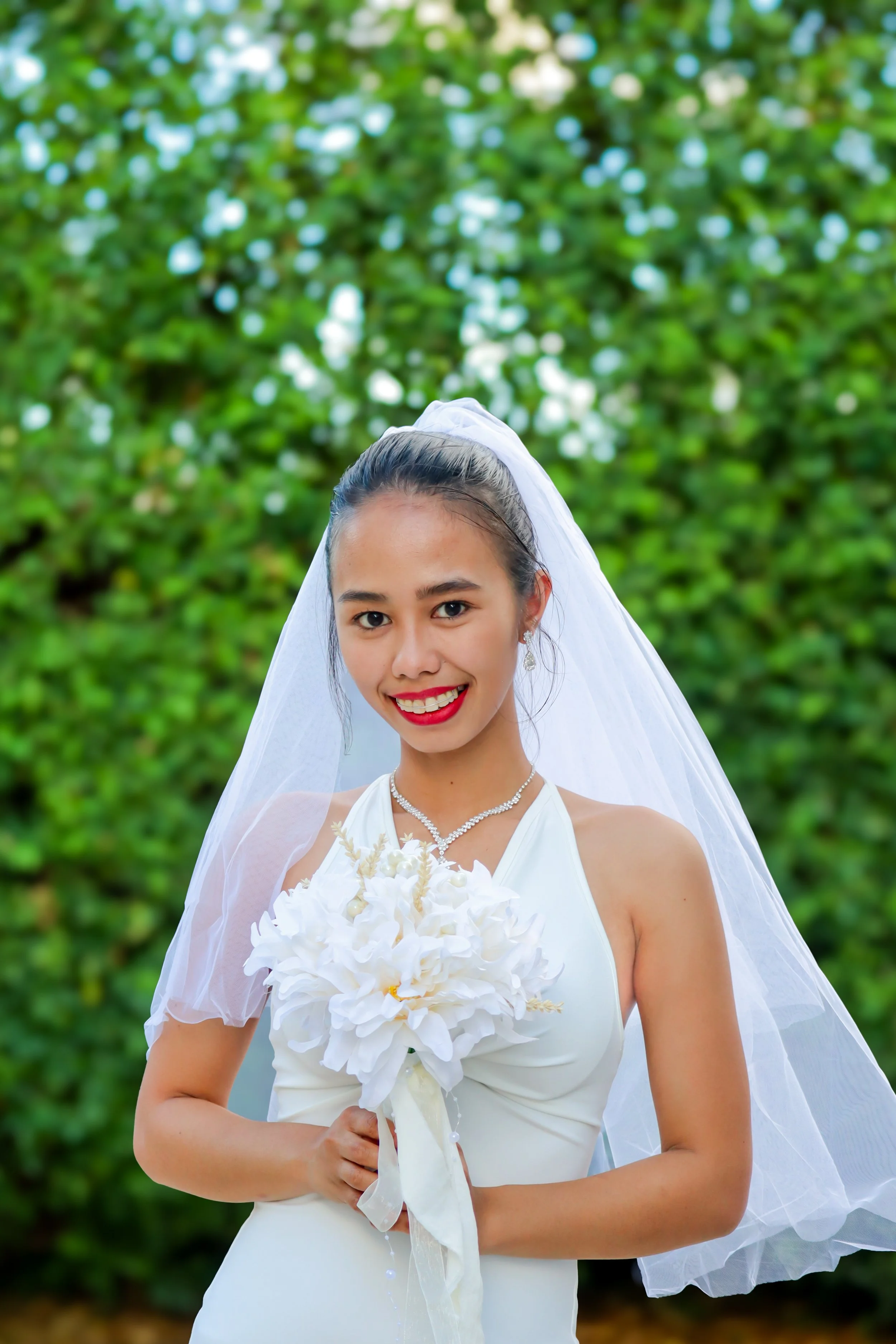 A smiling bride holding a bouquet of white flowers, wearing a white wedding dress and a veil, standing outdoors with green foliage in the background.