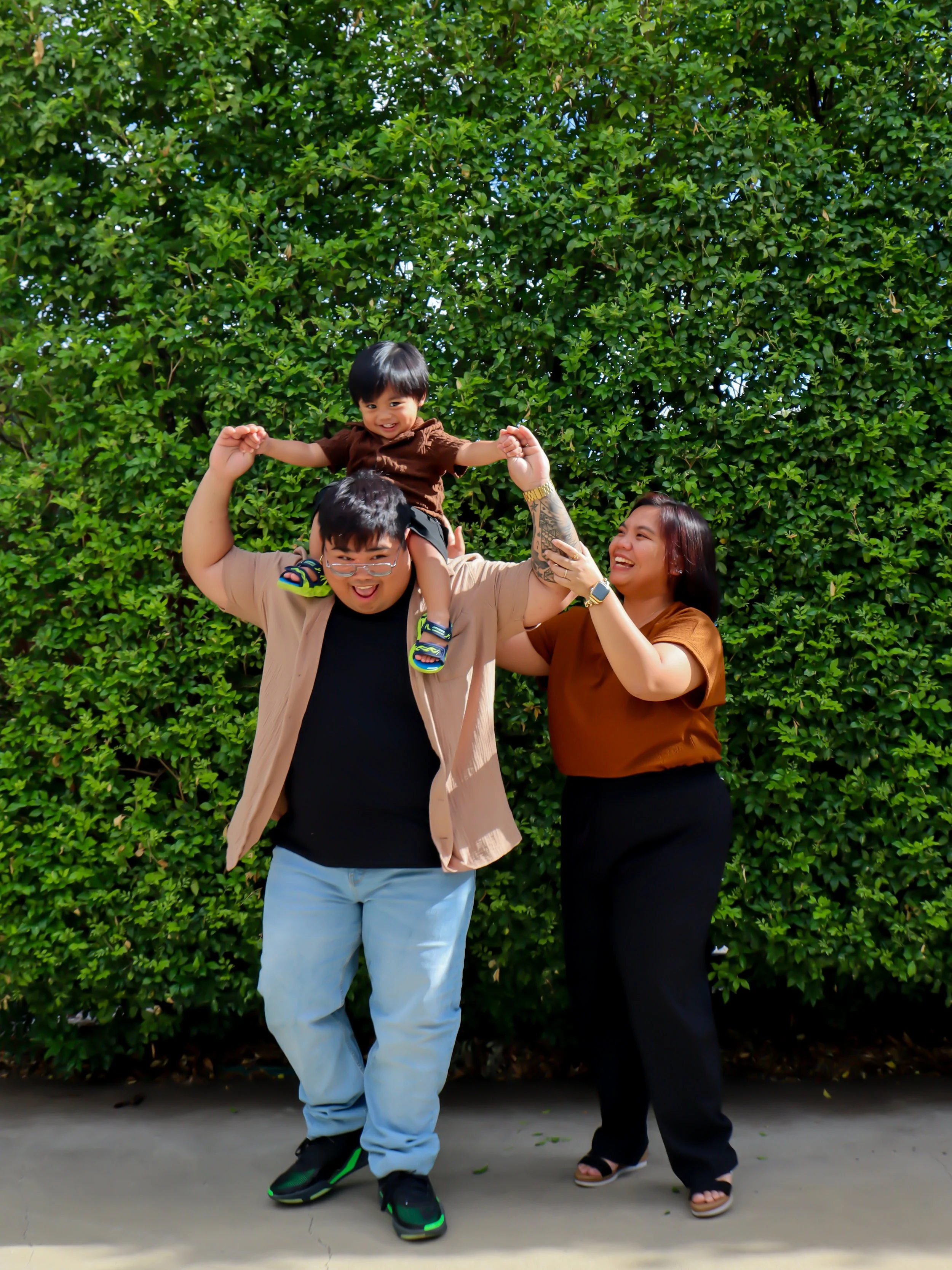 A family of three, including a man, woman, and young boy, enjoying time together outdoors in front of a green bush. The man is carrying the boy on his shoulders while the woman is smiling and reaching for the boy's arm. All are smiling and appear hap
