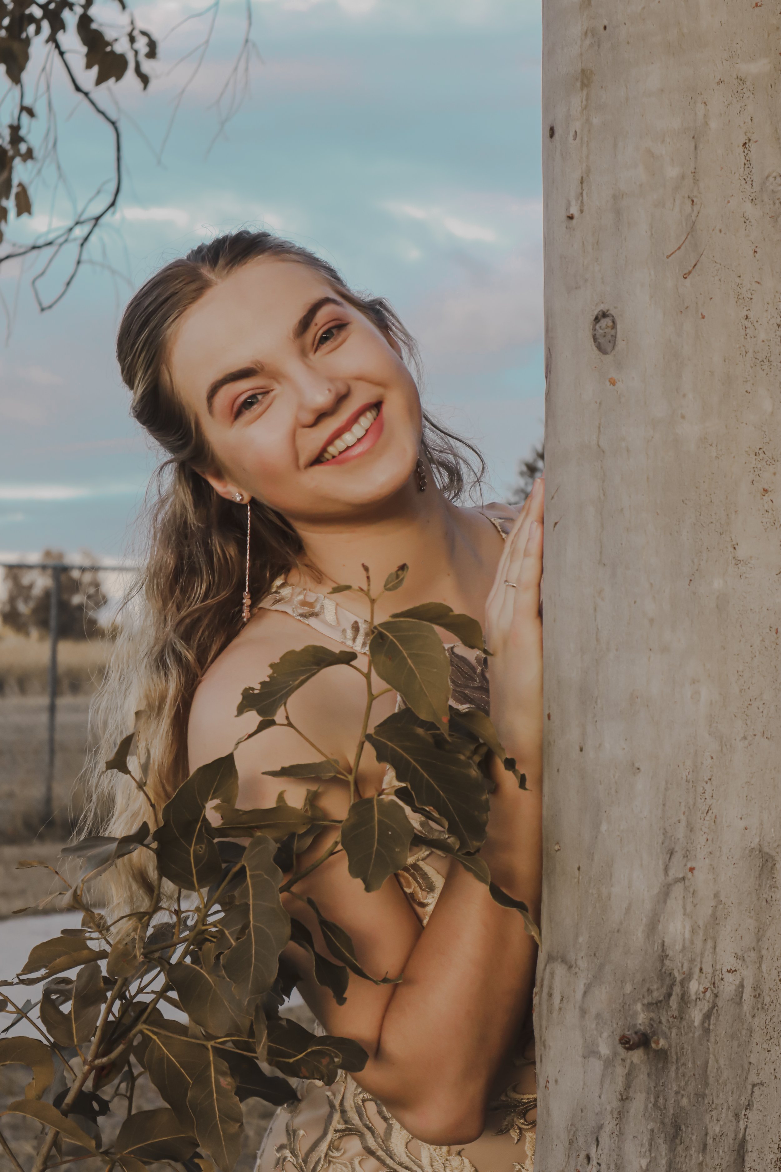 A young woman with long, wavy hair is smiling and peeking from behind a tree trunk outdoors during daylight. She has a nose piercing, earrings, and is wearing a sleeveless dress with a floral pattern. There are green leaves in the foreground, and a f