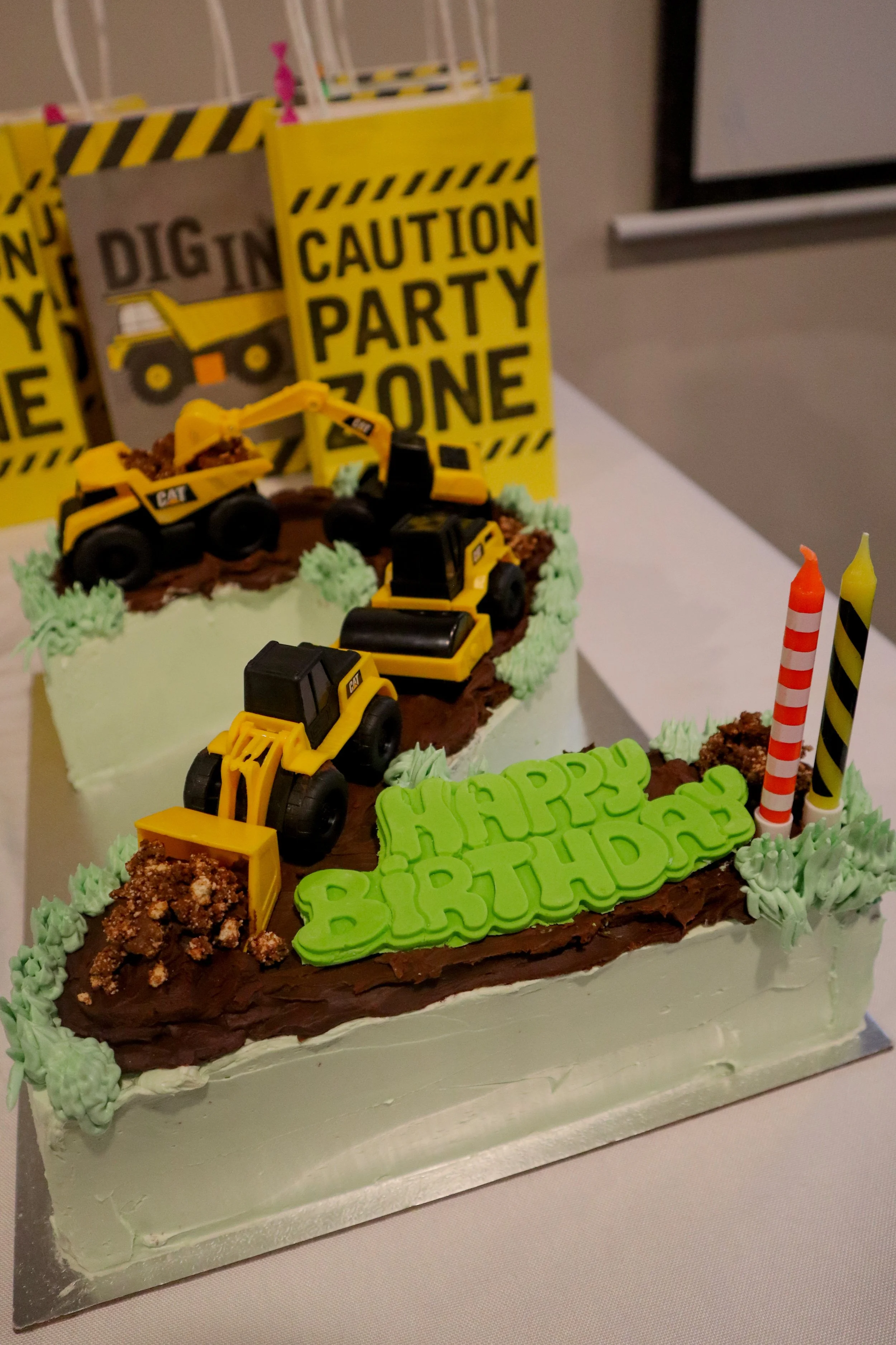 Construction-themed birthday cake decorated with toy construction vehicles, green frosting, and caution signs in the background reading "Caution Party Zone" and "Dig In". The cake also has a green "Happy Birthday" sign.
