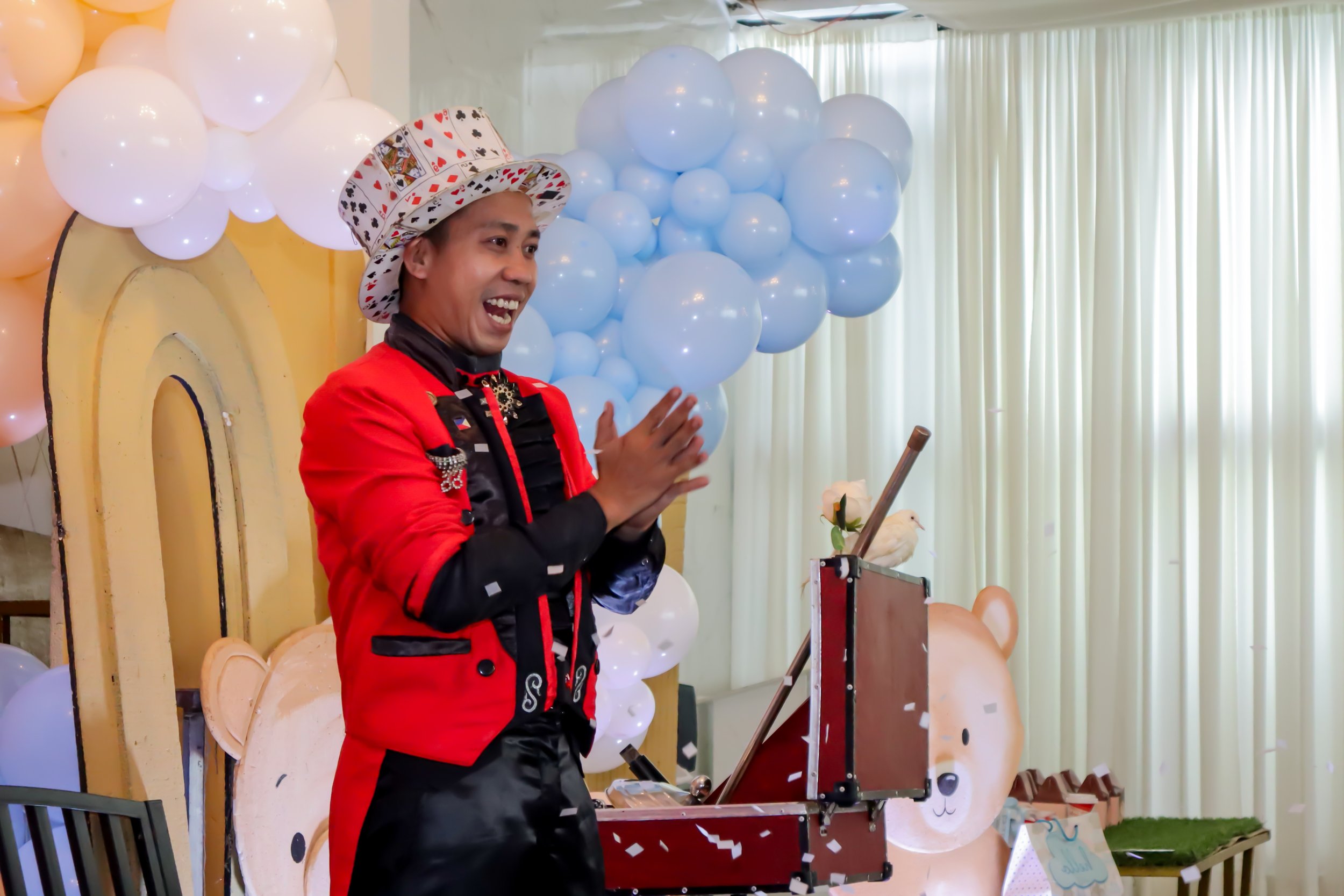 A magician in a red jacket, black shirt, and a casino-themed hat performing a magic trick in front of a decorated backdrop with balloons and teddy bear faces, with paper confetti falling around.