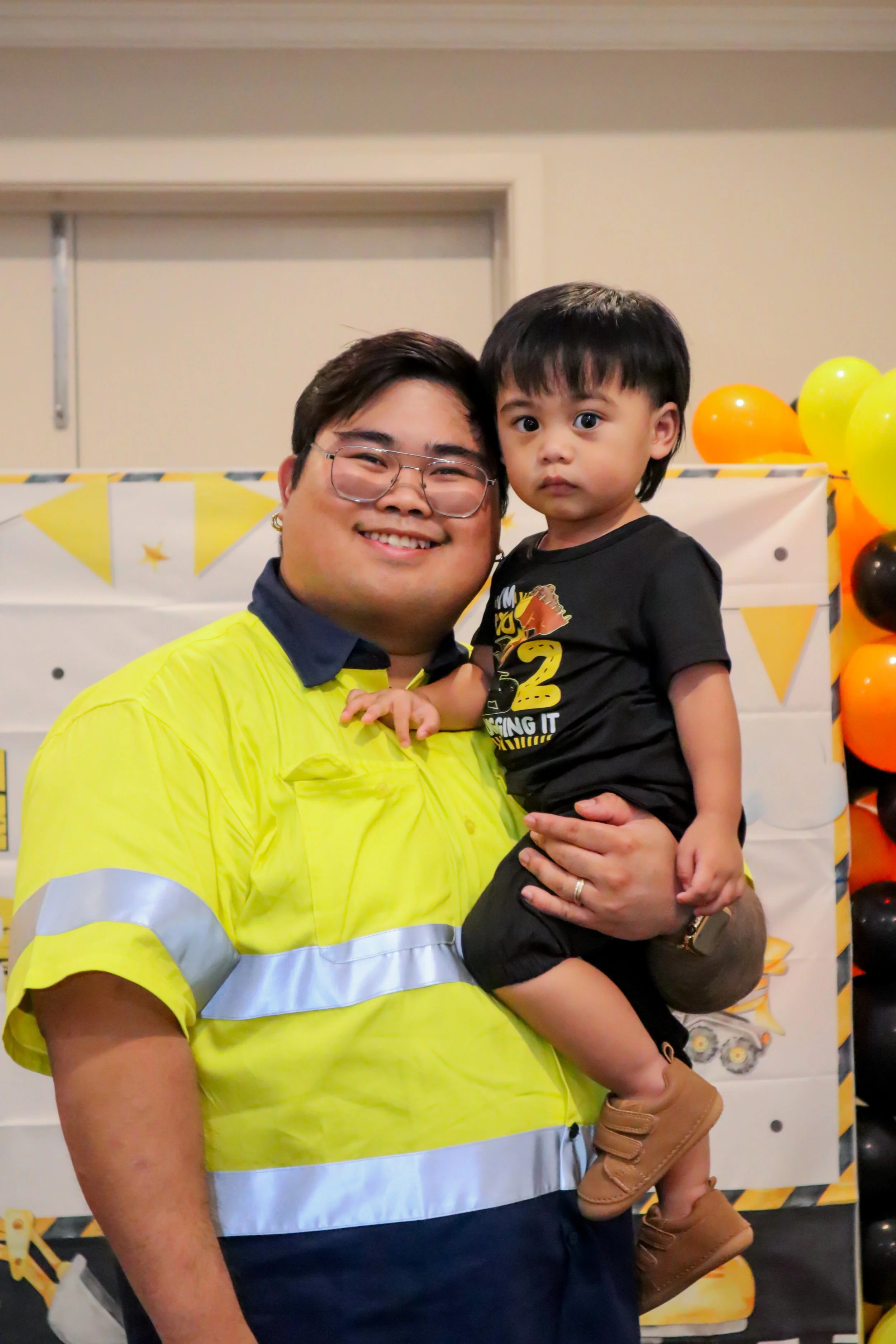 A smiling man in a bright yellow safety shirt holding a young boy in front of a decorated background with yellow and black balloons.
