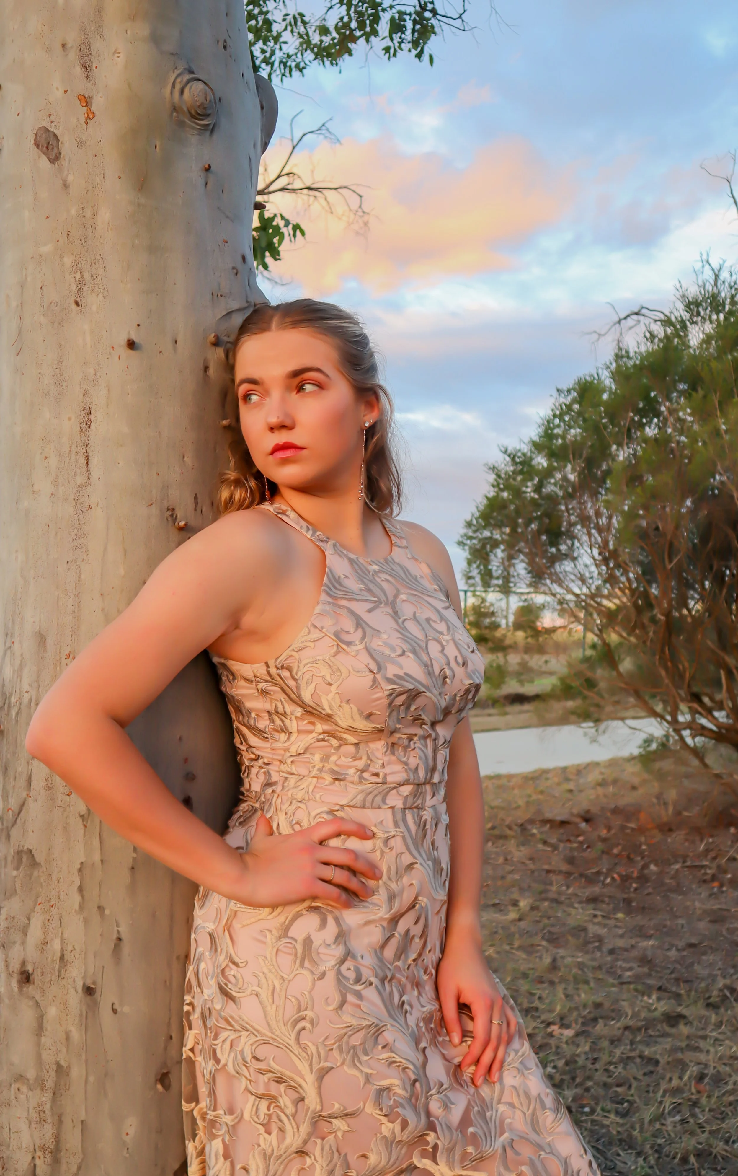 Young woman in a patterned dress leaning against a large tree trunk outdoors during sunset, with trees and a body of water in the background.