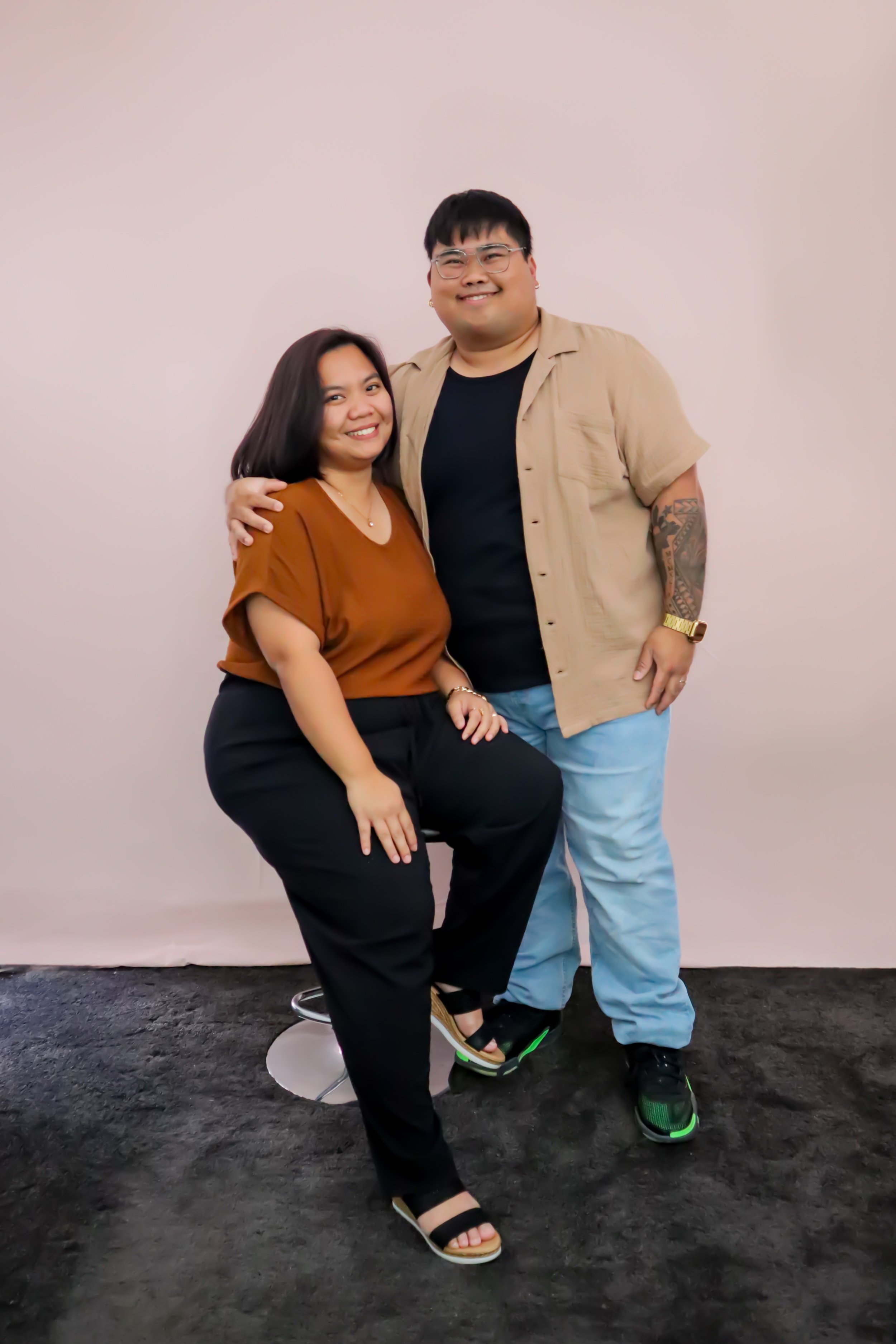A smiling woman sitting on a stool with a hand on her knee, and a smiling man standing next to her with his arm around her shoulders, both posing for a photo in front of a plain pink wall.