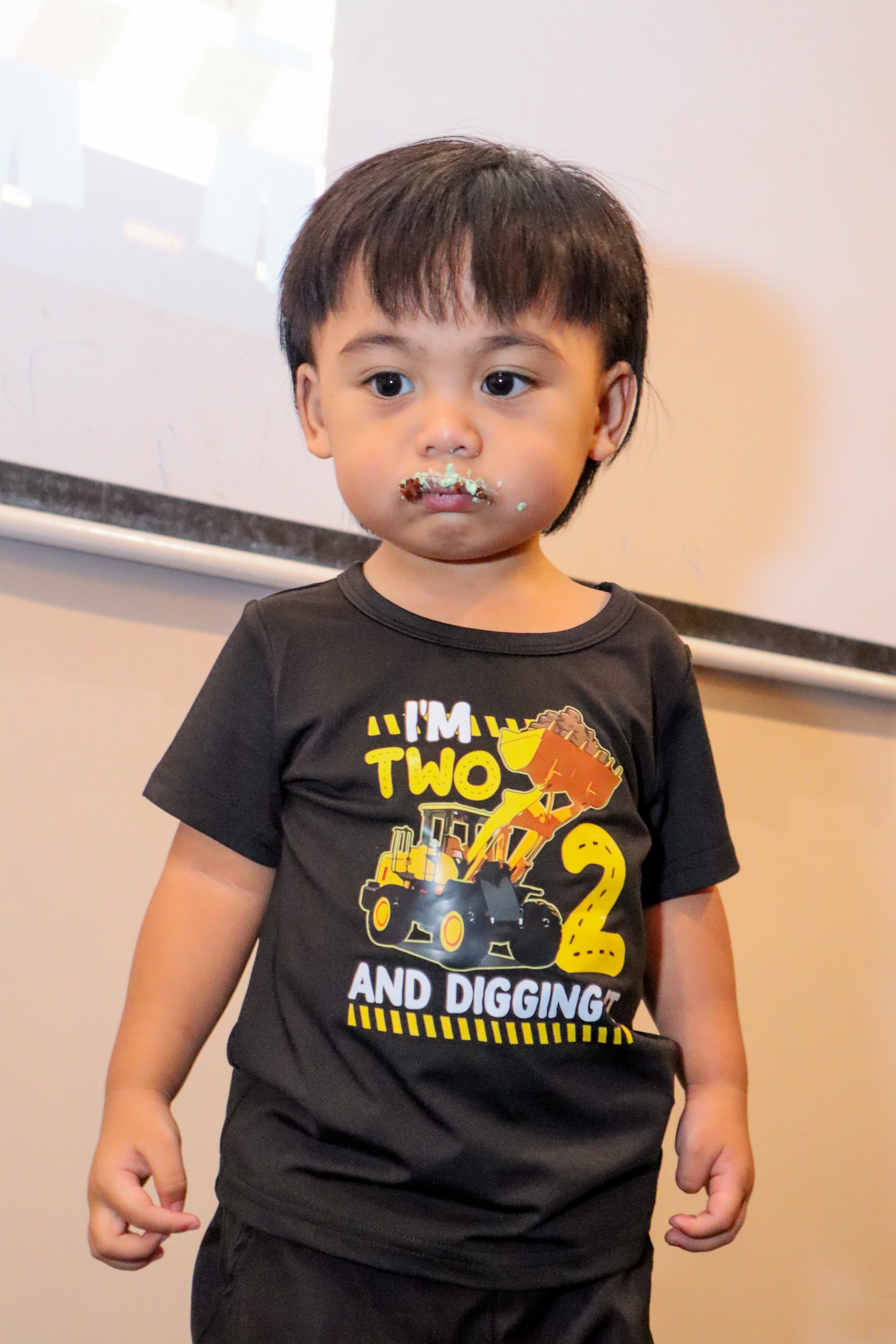 A young boy with dark hair and brown eyes, wearing a black T-shirt with a construction vehicle and the text 'I'M TWO AND DIGGING,' stands indoors with a messy face and cake or food around his mouth.