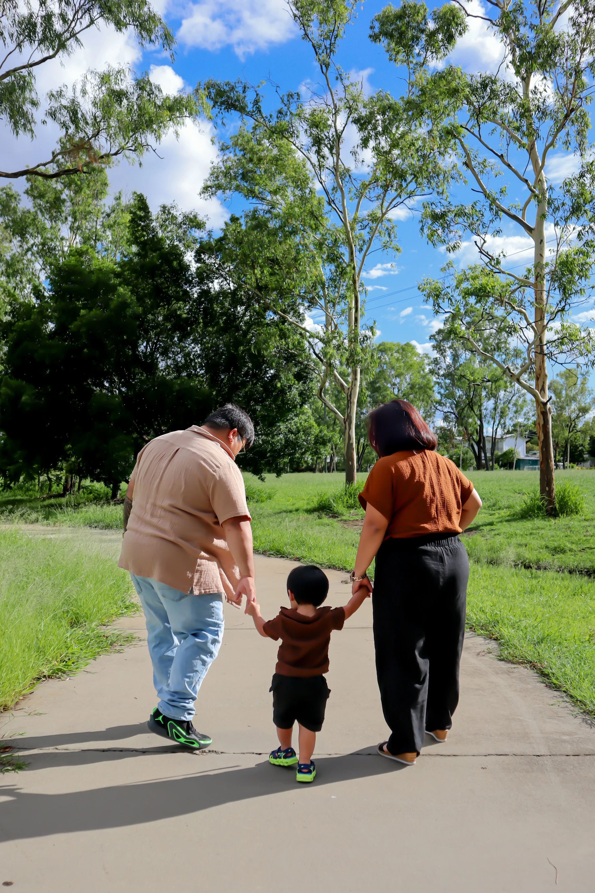 A young boy is walking hand-in-hand with two adults, a man and a woman, on a paved path in a park with green grass and tall trees, under a partly cloudy sky.