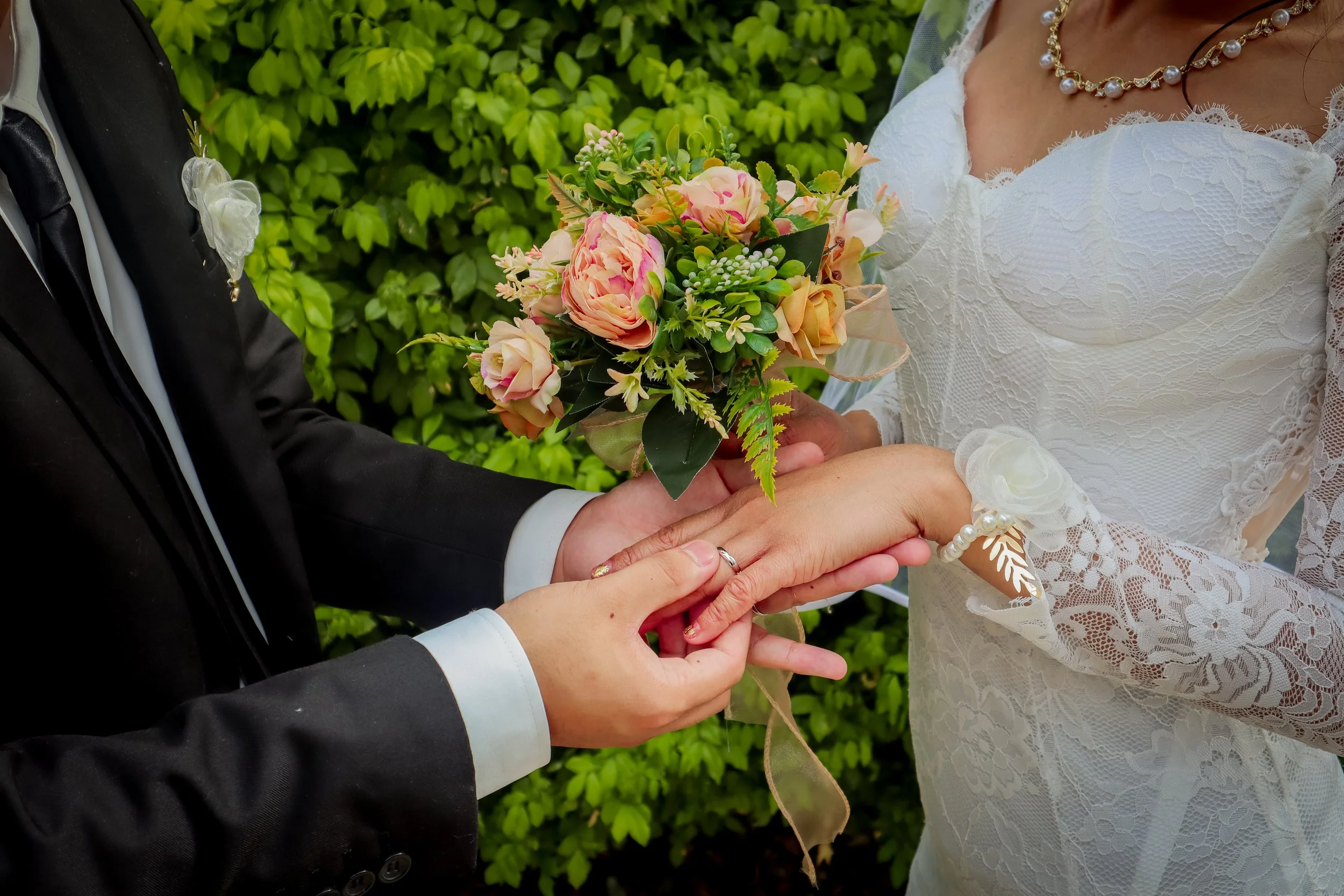 A bride and groom exchanging rings outdoors with greenery in the background. The groom is wearing a black tuxedo, and the bride is wearing a white lace wedding dress with jewelry and holding a bouquet.