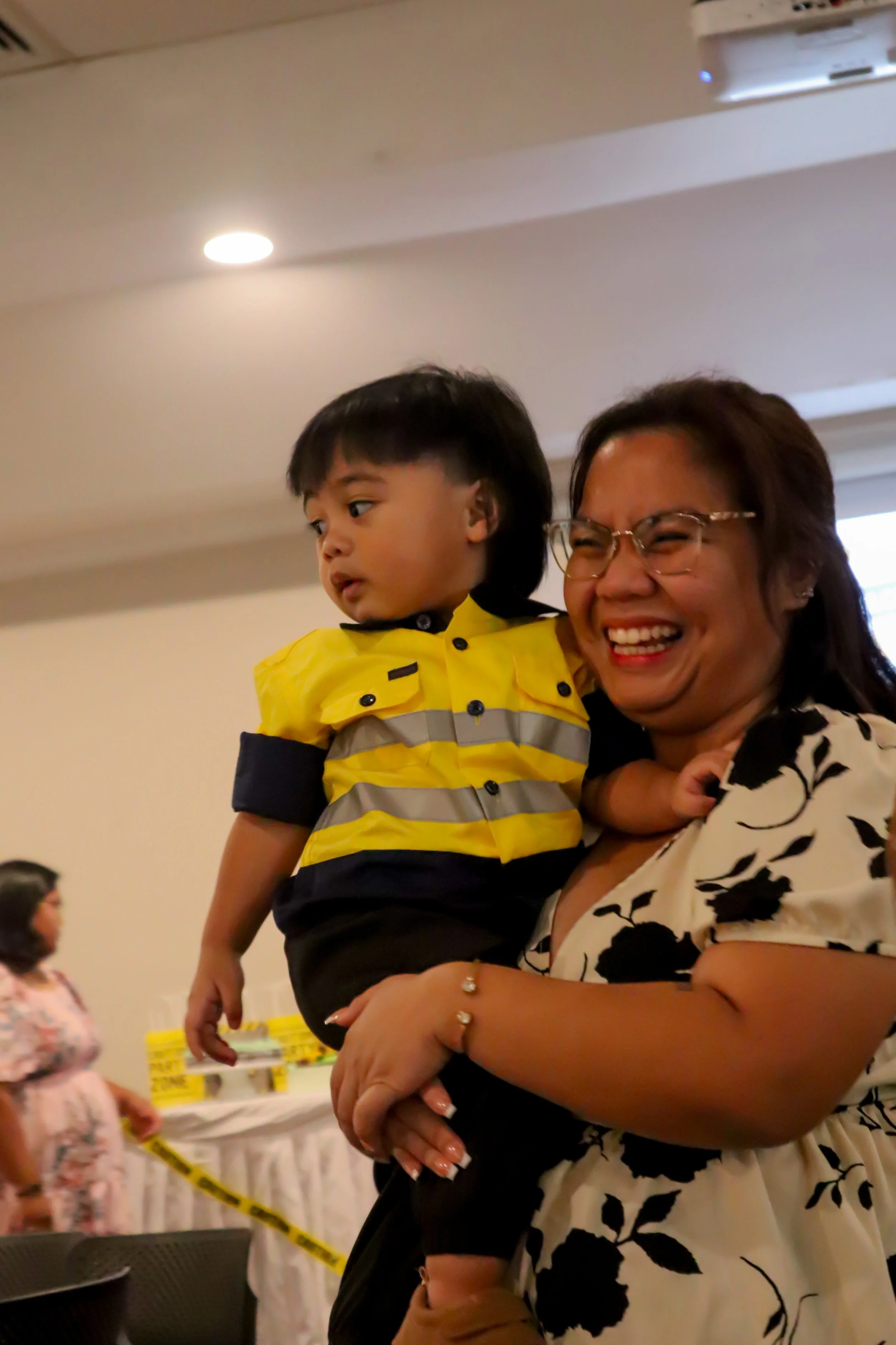 A woman with glasses and a woman in a patterned blouse holding a young boy in a yellow and black shirt, smiling and laughing at an indoor event.