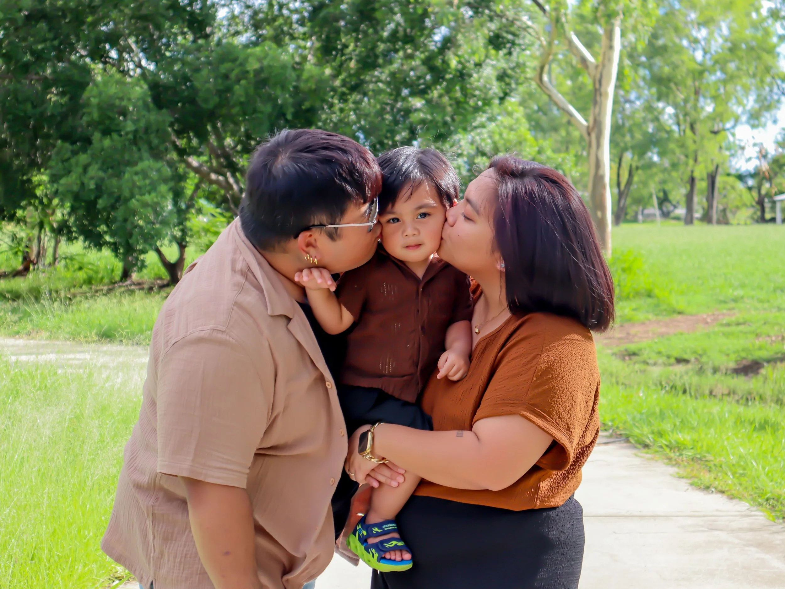 A loving family of three, with a young boy in the middle, being kissed on each cheek by his mother and father in a park with green trees and grass.