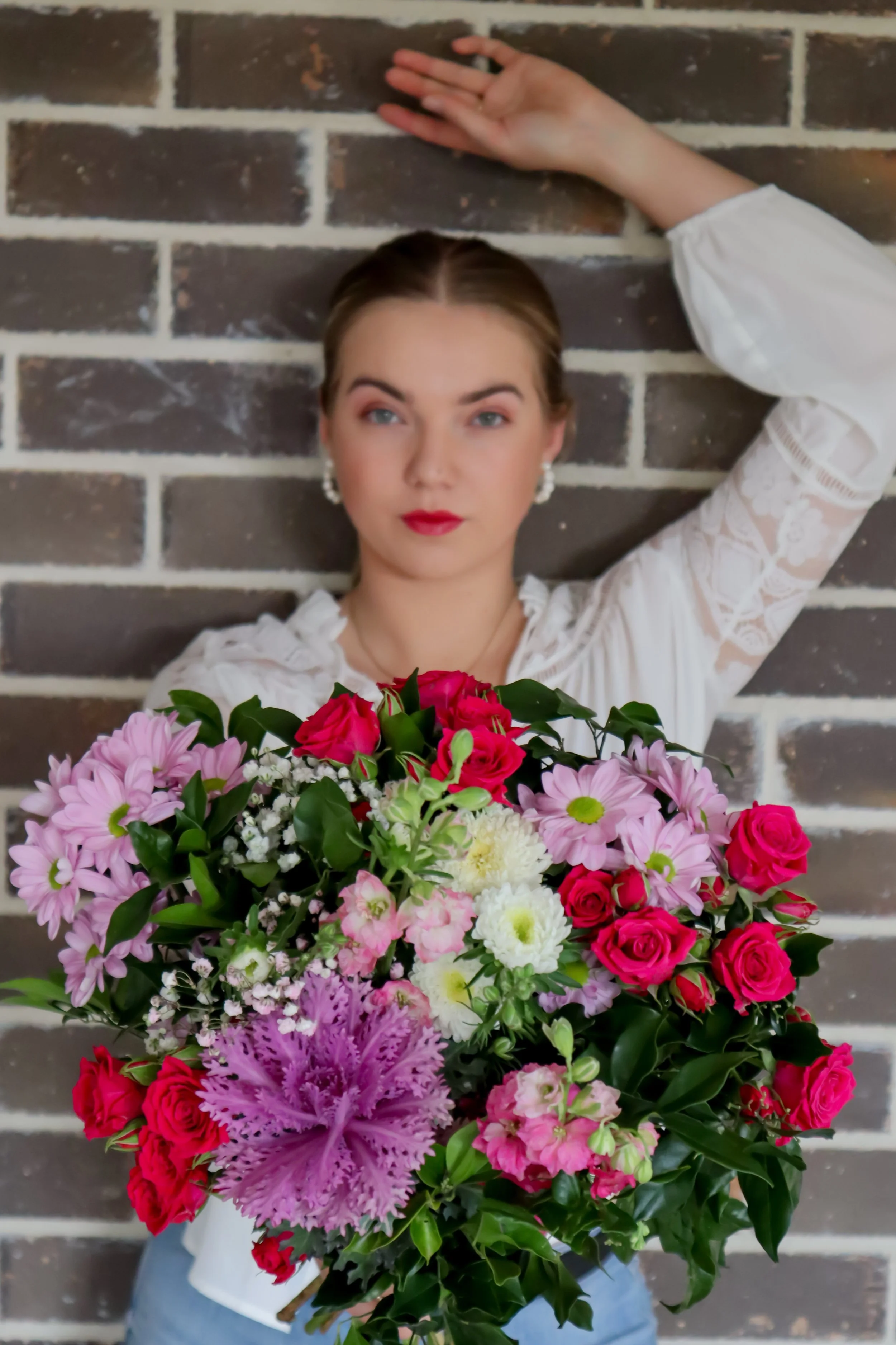 A woman with fair skin, makeup, and earrings holds a large bouquet of pink, red, white, and purple flowers, standing against a brick wall background.