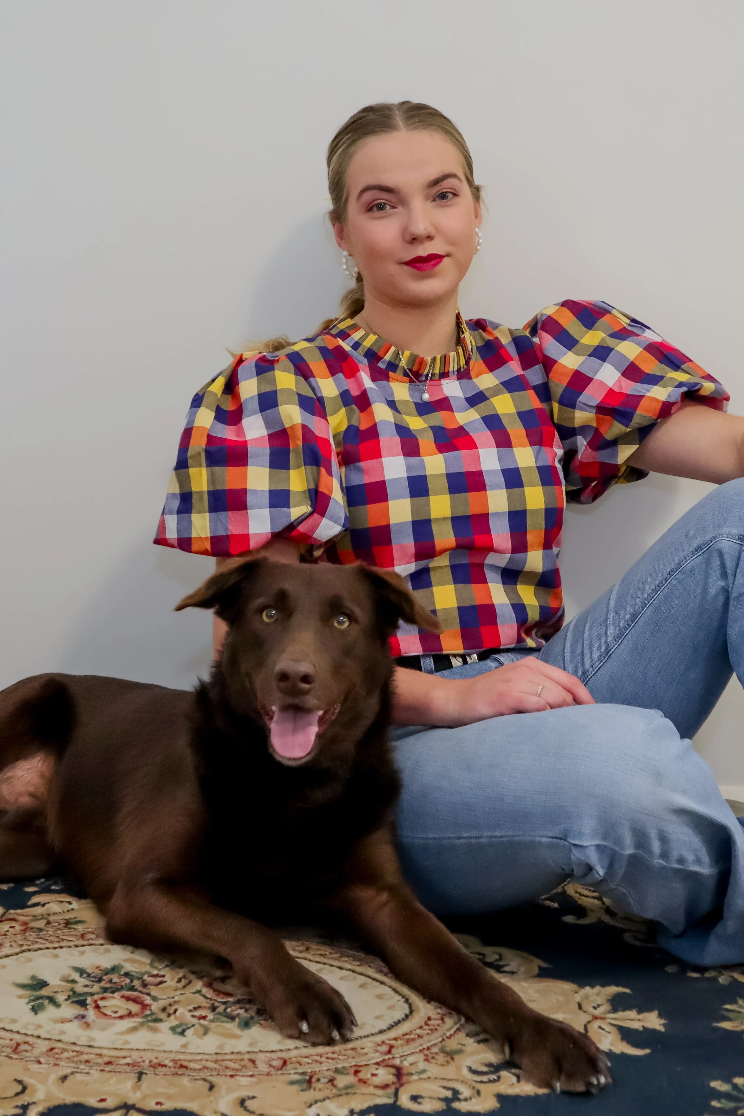 Young woman sitting on the floor with a brown dog lying beside her. The woman has blonde hair in a braid, is wearing a colorful checkered puff-sleeve shirt and blue jeans. The dog has a happy expression, tongue out, and is looking at the camera. They
