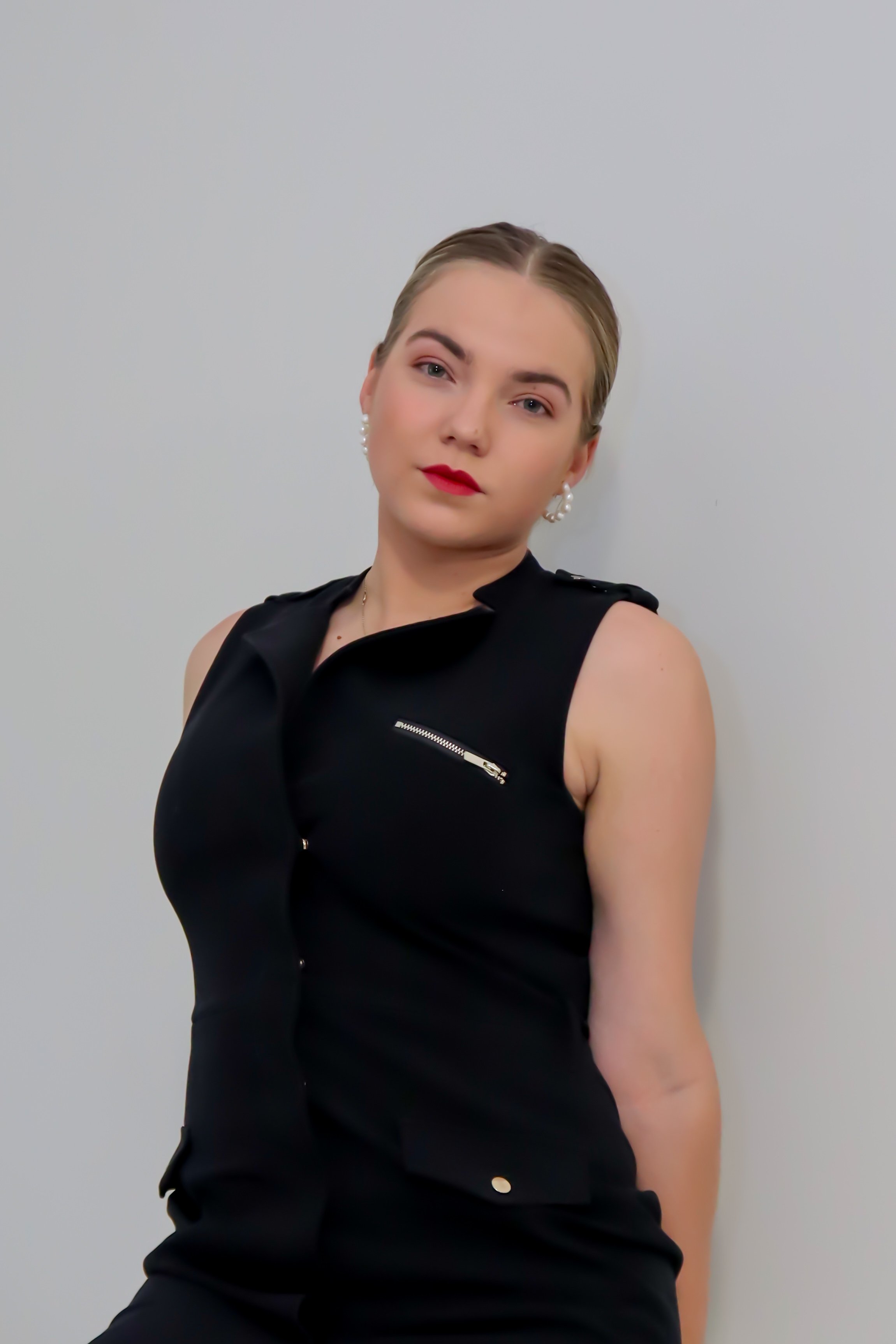 A young woman in a black sleeveless top with a zipper detail and pearl earrings stands against a plain background, looking into the camera with a neutral expression.