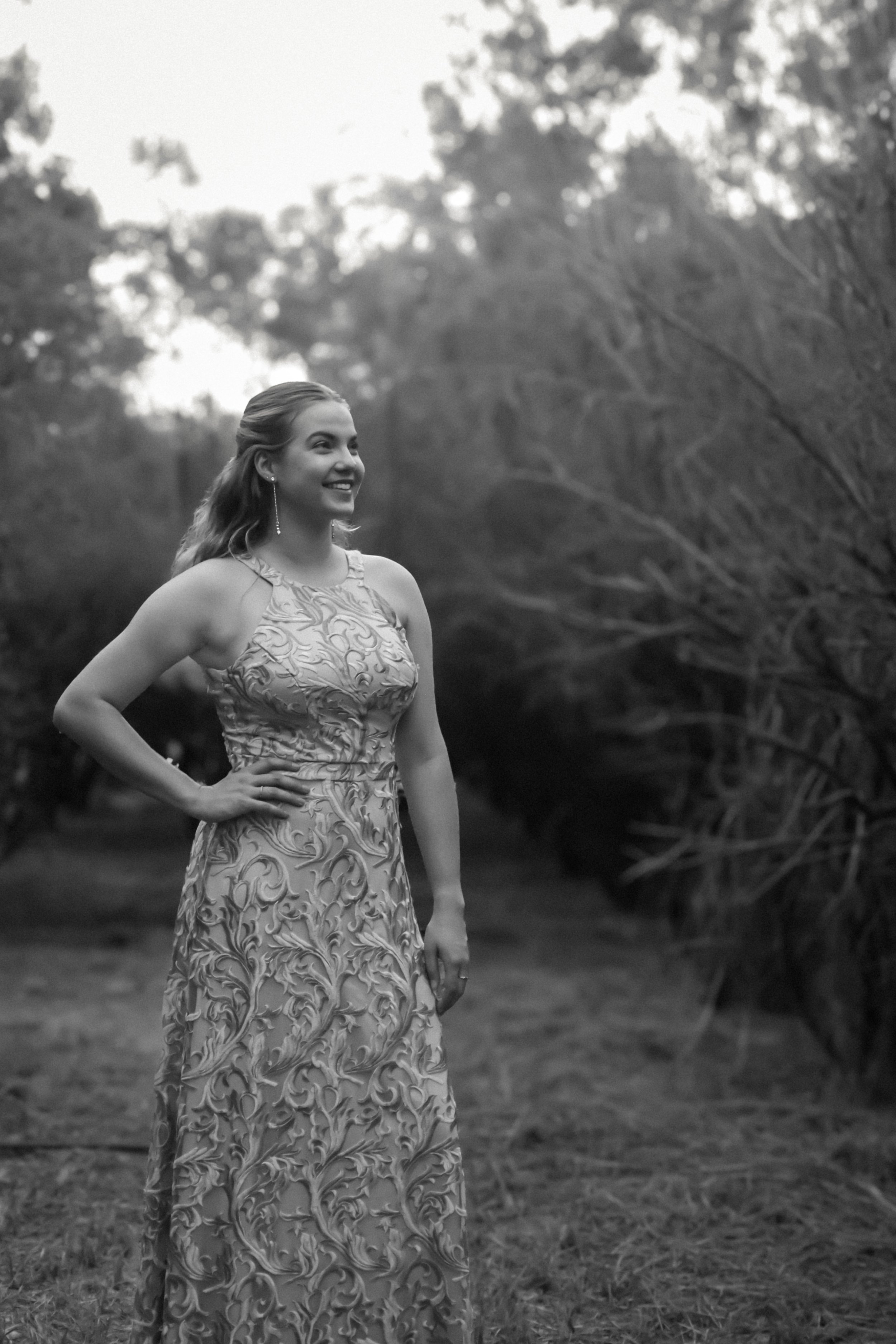 A young woman in an ornate sleeveless dress outdoors, smiling and looking to the side, with trees and a grassy area in the background, in black and white.