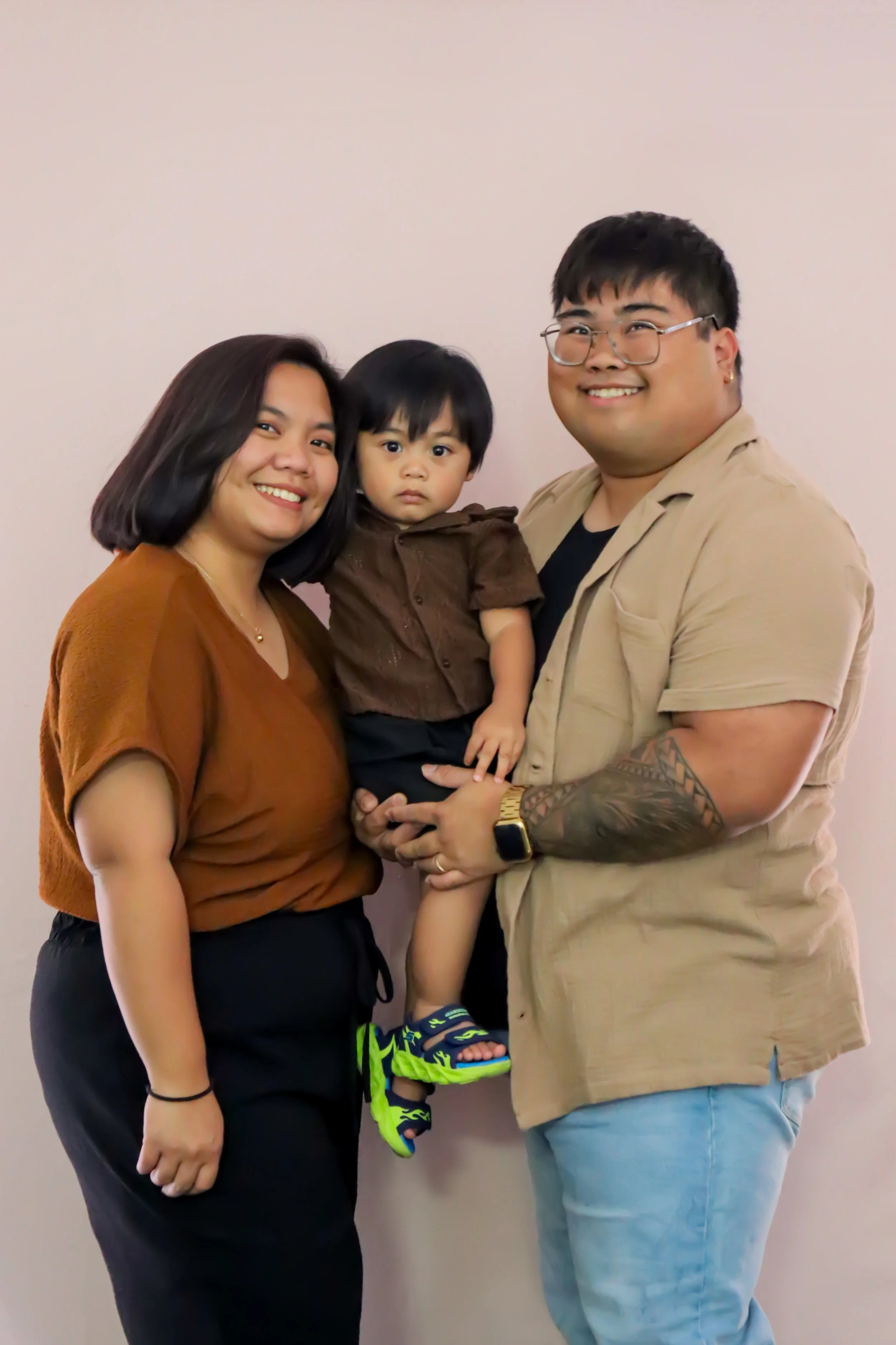 A family of three, a woman, a man, and a young boy, is standing against a plain light-colored wall, smiling at the camera. The woman has shoulder-length dark hair, wearing a brown top. The man has short black hair, glasses, a tan short-sleeved shirt,