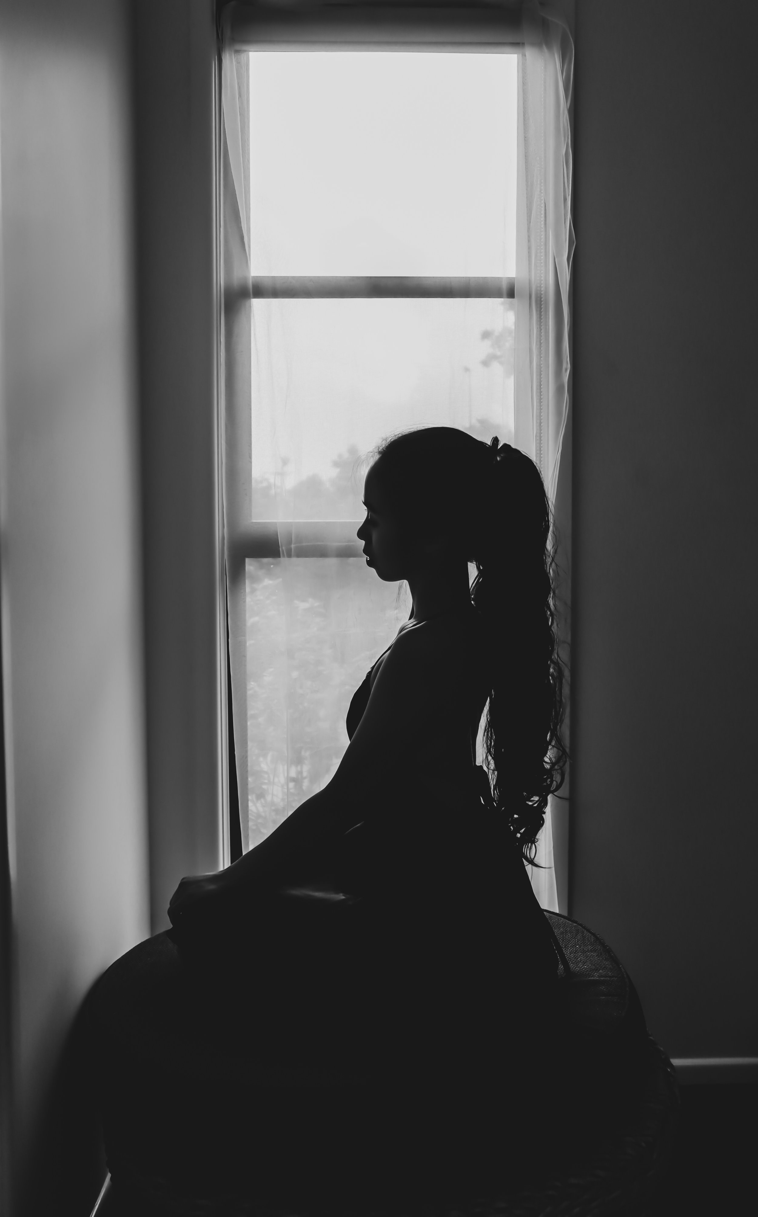 Silhouette of a woman with long hair sitting on a ball in front of a window with sheer curtains, with natural light coming through, in black and white.