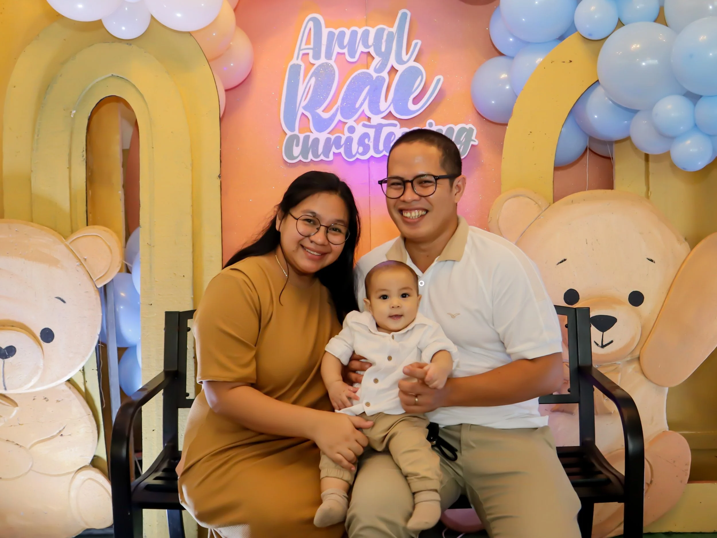A family of three, including a woman, a man, and a baby, sitting on a bench at a birthday celebration with a teddy bear and balloon decorations in the background.
