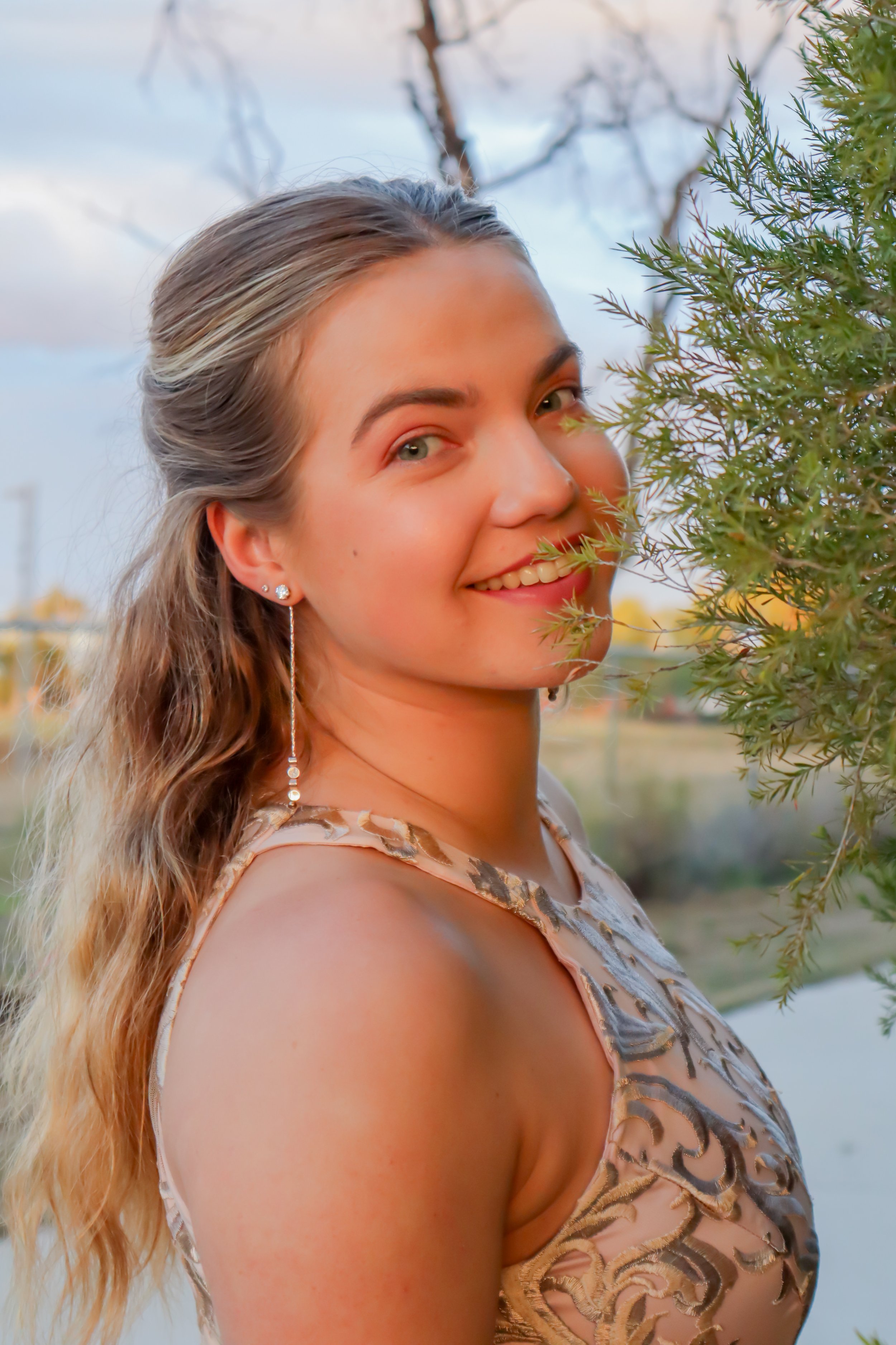 A young woman with wavy blonde hair, wearing a sleeveless dress with a gold pattern, smiling outdoors near a green leafy bush, during the daytime.