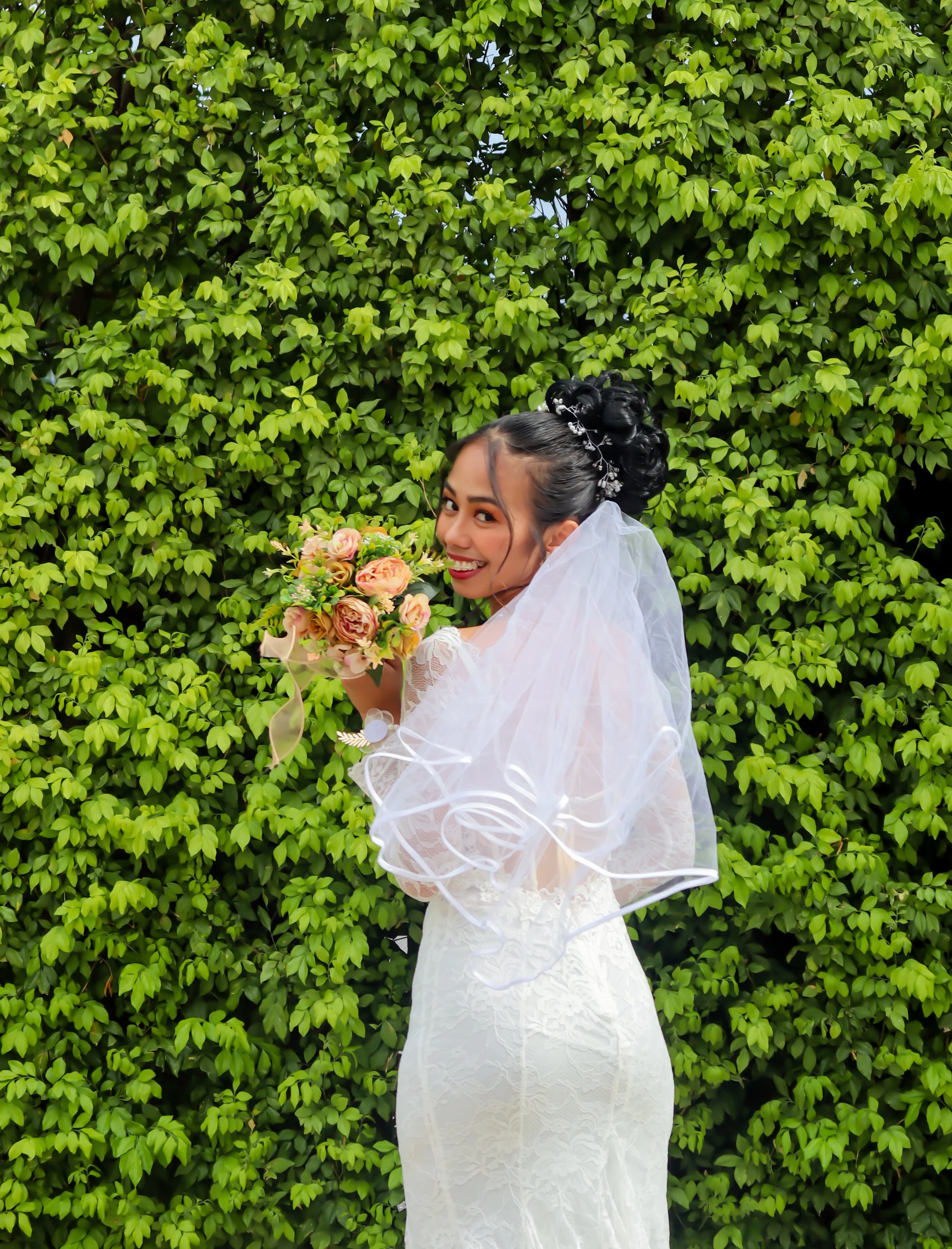 Bride in a white lace wedding dress holding a bouquet of pink and peach roses, smiling, standing in front of a lush green hedge.