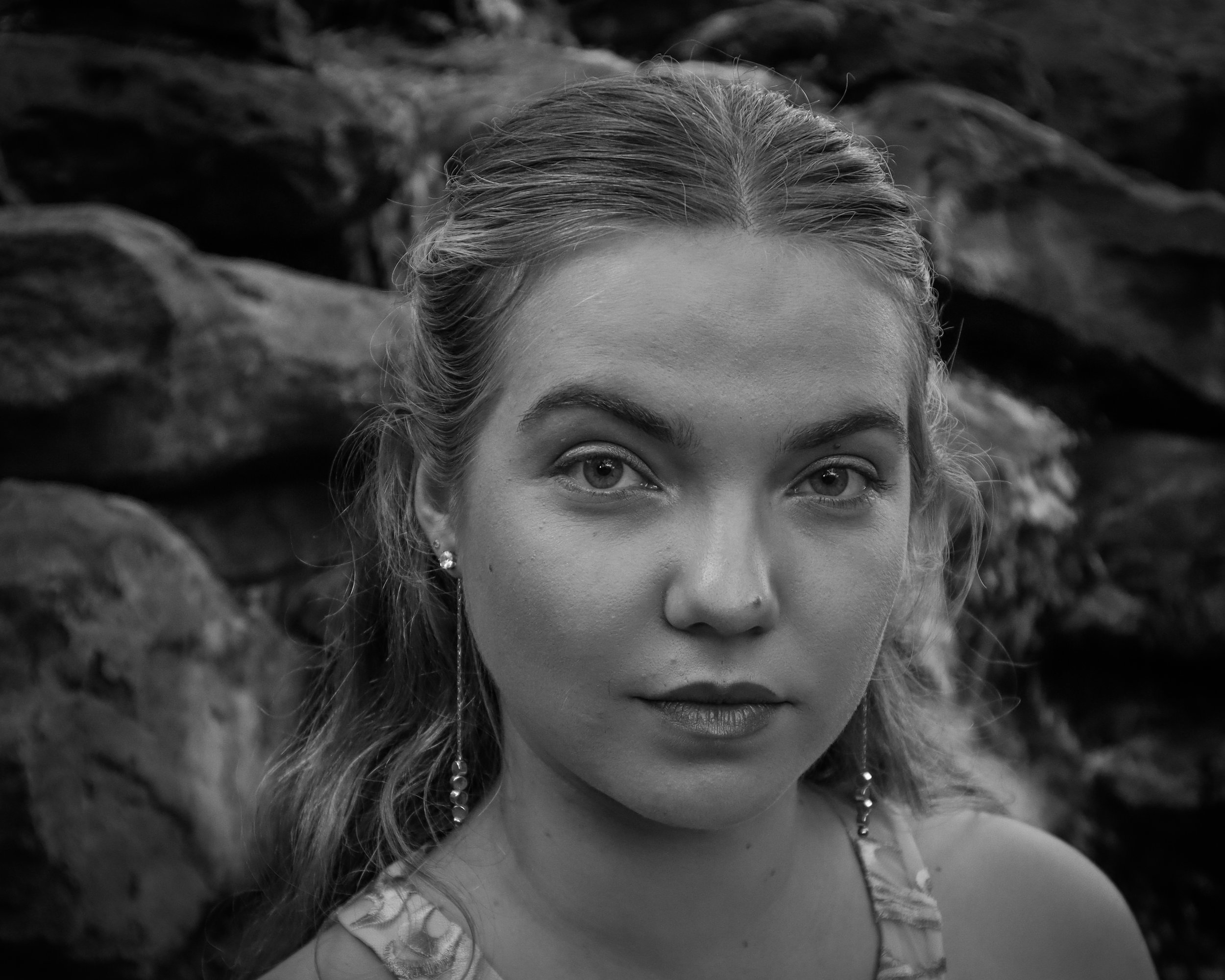 A close-up black and white portrait of a young woman with light-colored eyes, wearing earrings, and a floral-patterned top, standing in front of a background of large rocks.