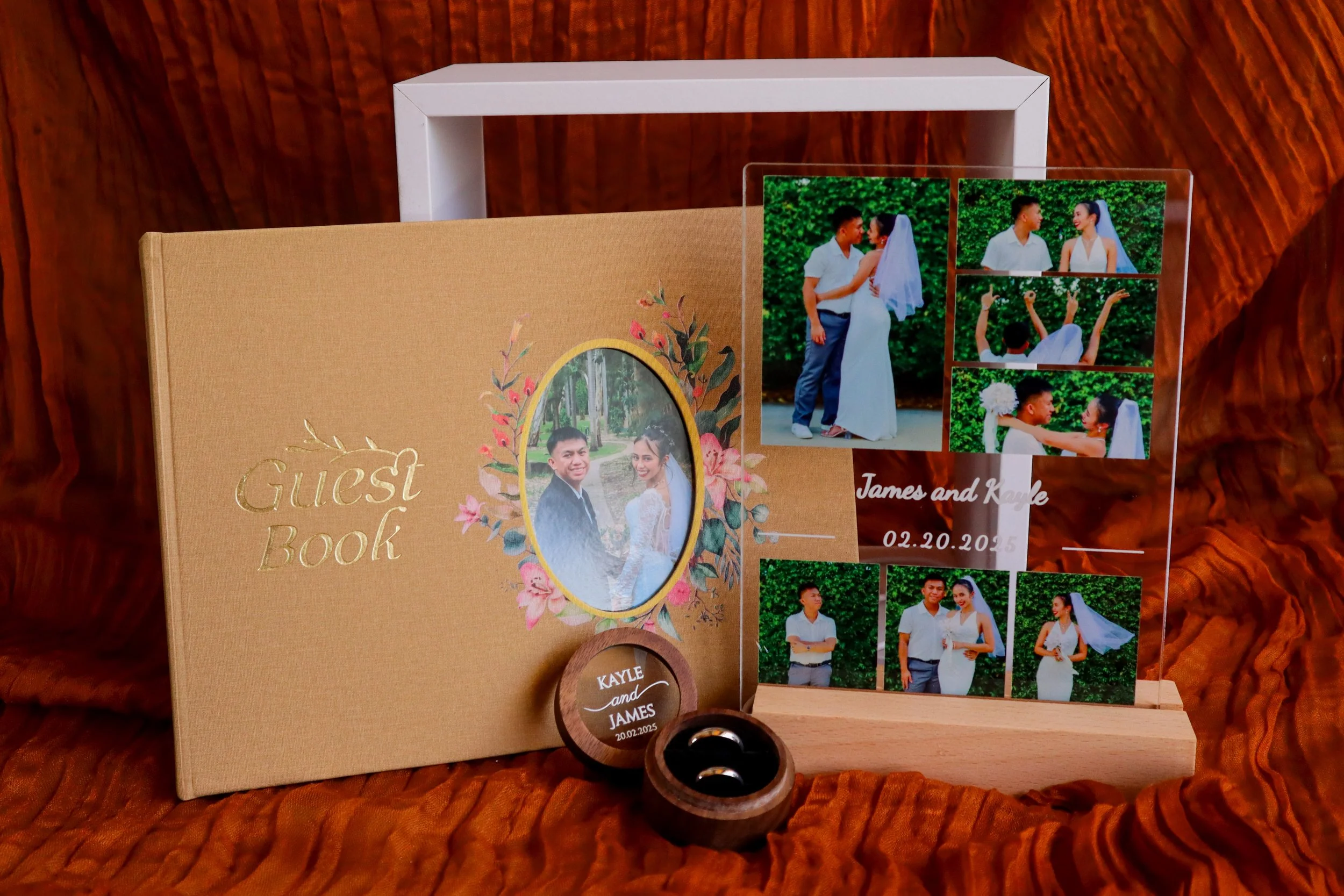 A wedding guest book with a floral cover, a photo of a couple in a forest, a clear acrylic stand with multiple wedding photos, a wooden ring box with engraved wedding details, and two wedding rings on a textured orange fabric background.