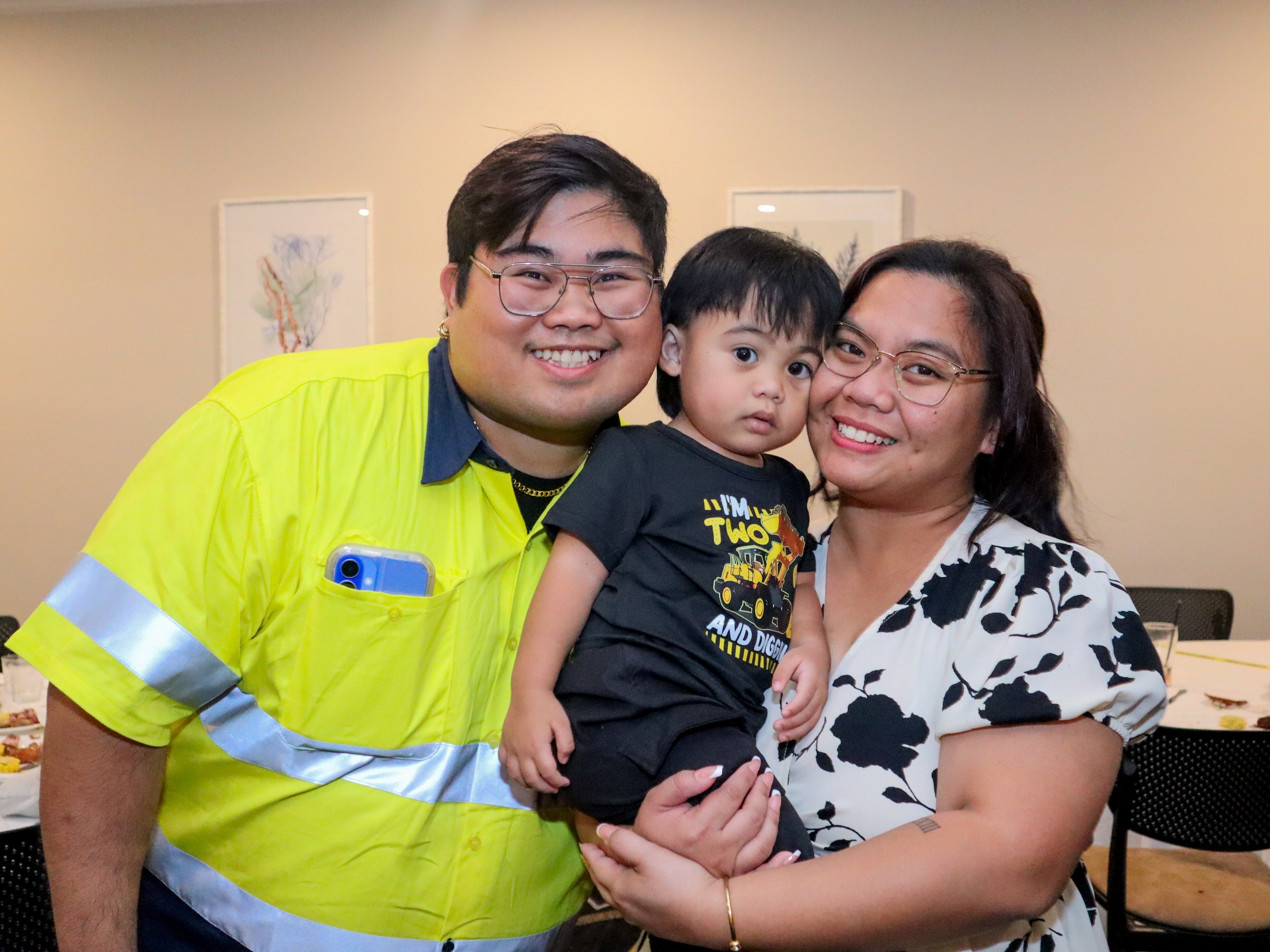 A family of three smiling and posing for a photo indoors. The father is wearing a bright yellow shirt with reflective stripes, the mother is wearing a white and black floral dress, and the young son is sitting on the mother's arm wearing a black t-sh