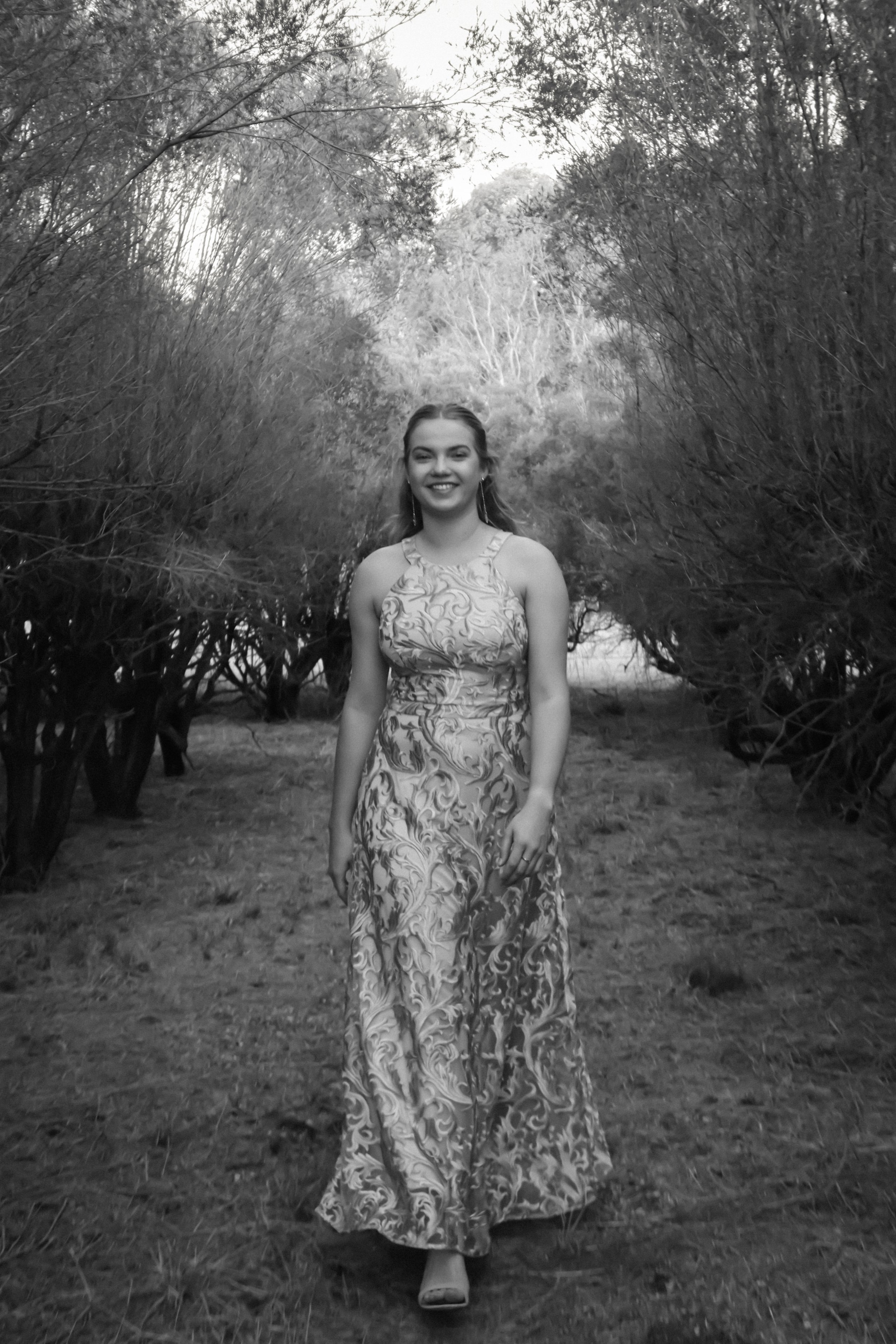 A smiling woman in a patterned dress walking down a wooded path in black and white