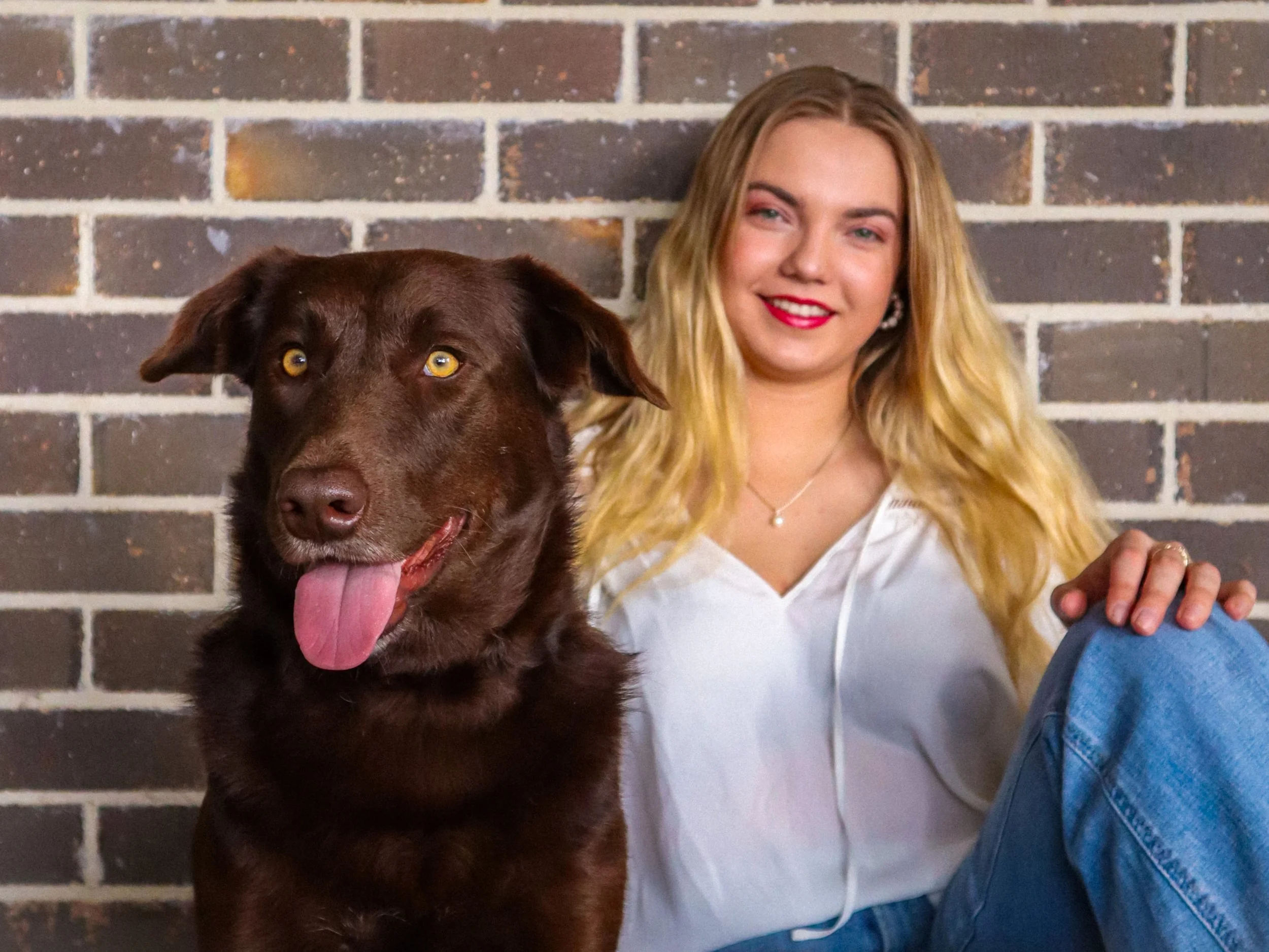 A woman with blonde hair and wearing a white top sits against a brick wall, next to a chocolate Labrador retriever with yellow eyes and its tongue out.