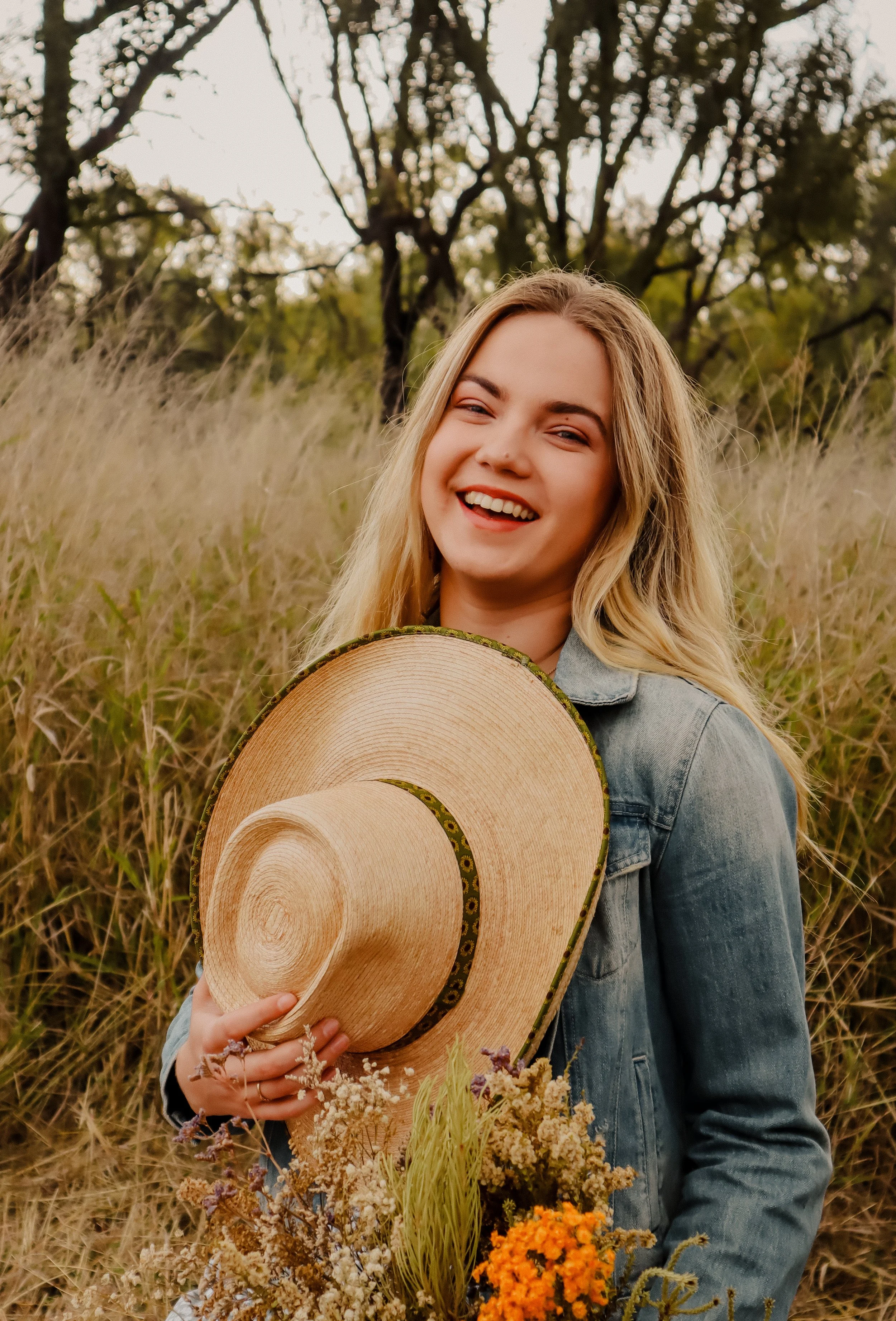 A young woman with long blonde hair, smiling, holding a woven straw hat and a bouquet of wildflowers in a grassy field with trees in the background.