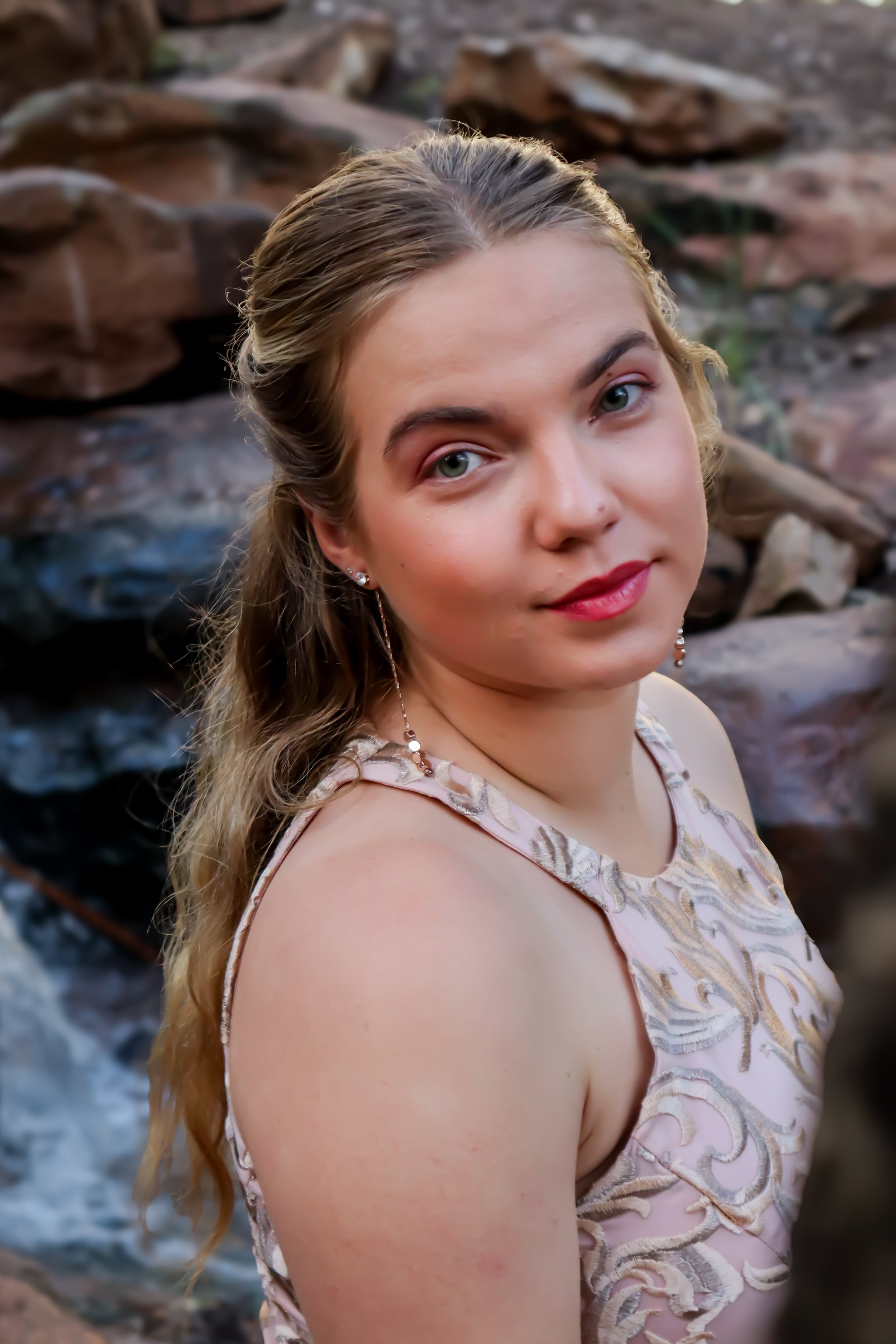 A young woman with long, wavy hair and blue eyes, wearing a sleeveless, embroidered dress with gold and beige patterns. She has earrings and a necklace, and stands in front of a rocky background, looking directly at the camera with a slight smile.