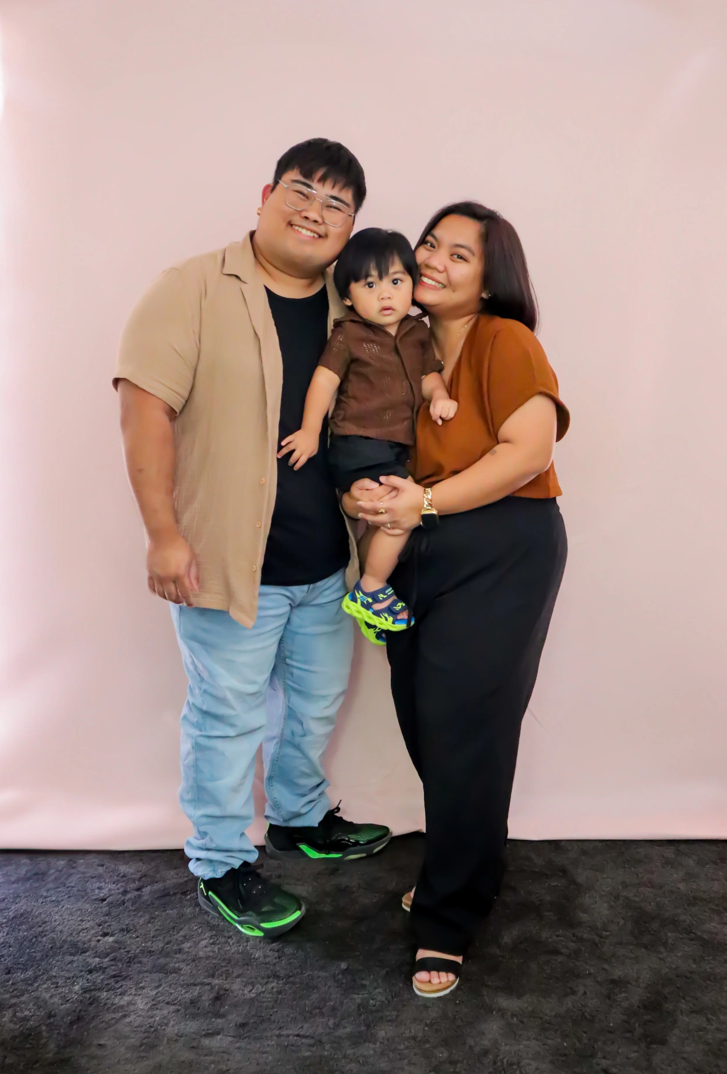 A smiling family of three standing against a plain background. The father wears glasses, a beige short-sleeve shirt, and light blue jeans. The mother has shoulder-length dark hair, a brown top, and black pants. They are holding a young boy wearing a 