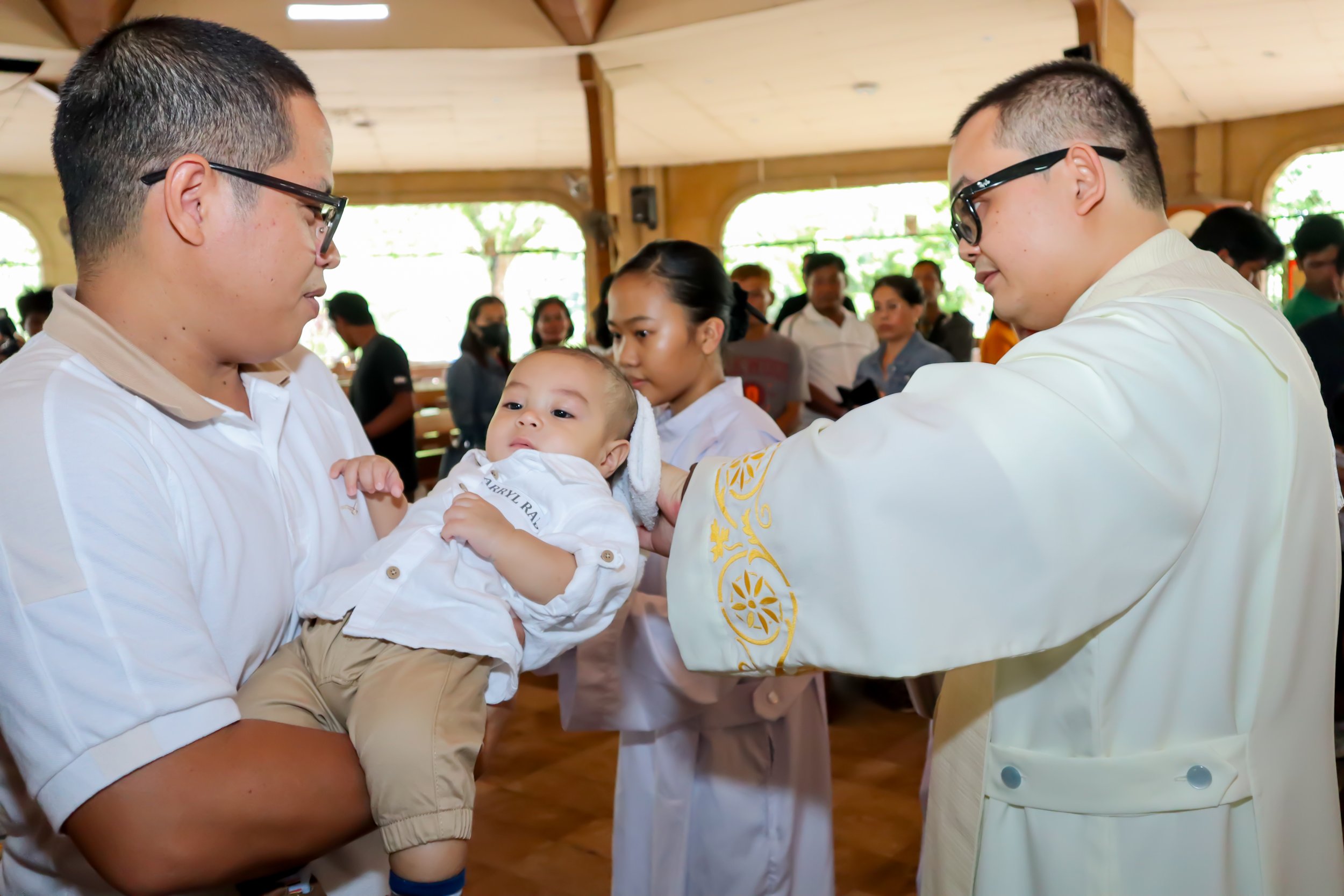 A baby being baptized held by an adult, with a priest performing the baptism in a church with many people in attendance.