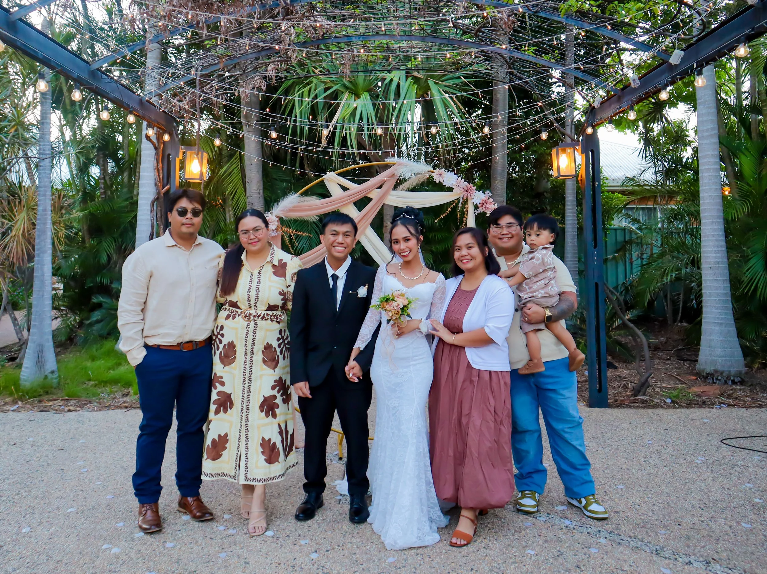 Group of seven people, including a bride in a white wedding dress and a groom in a black suit, posing outdoors under string lights and decorative fabric draping, with trees and plants in the background.