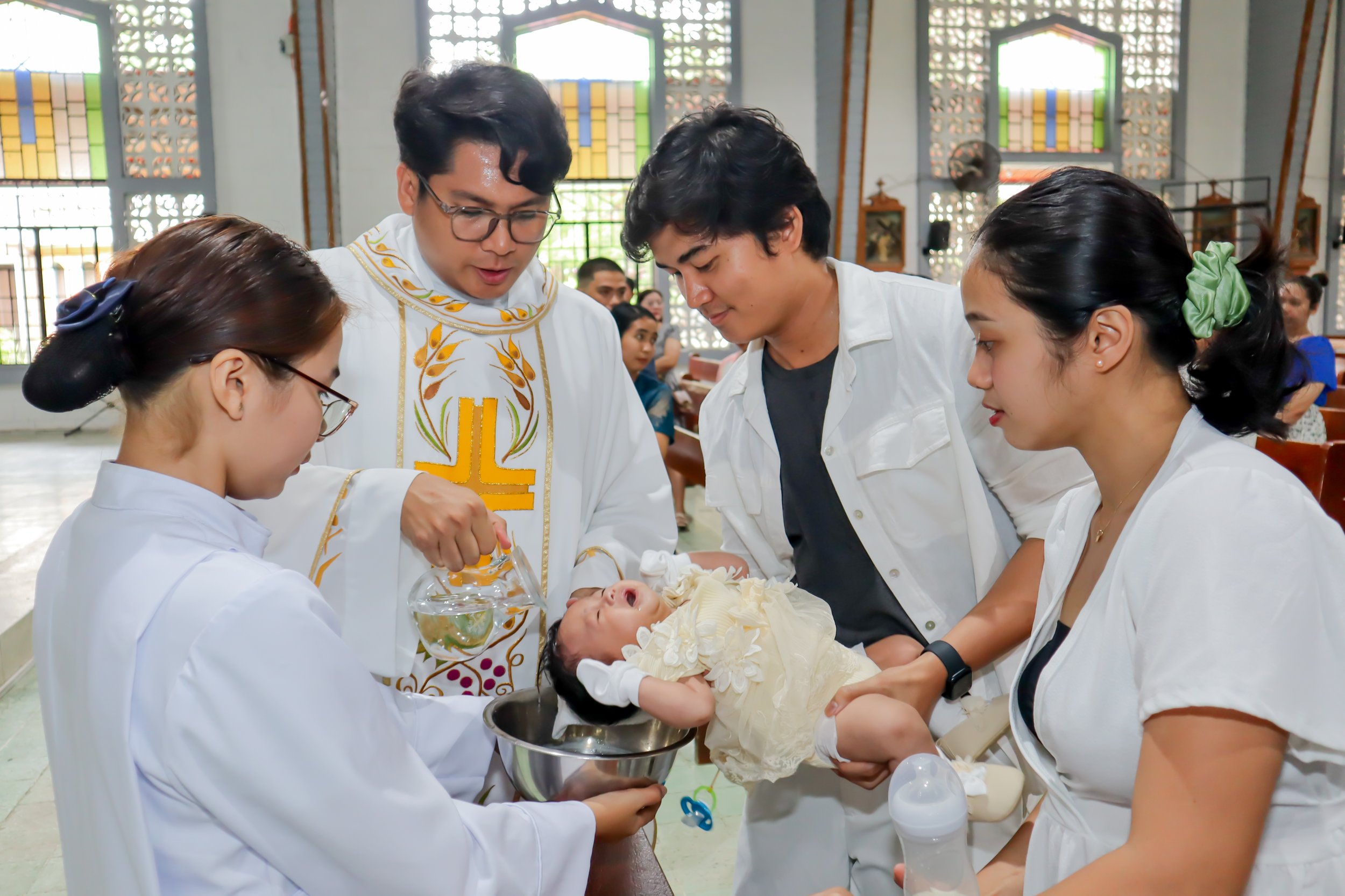 A group of people, including a priest, a nurse, and family members, participate in a baptism ceremony for a baby in a church setting.