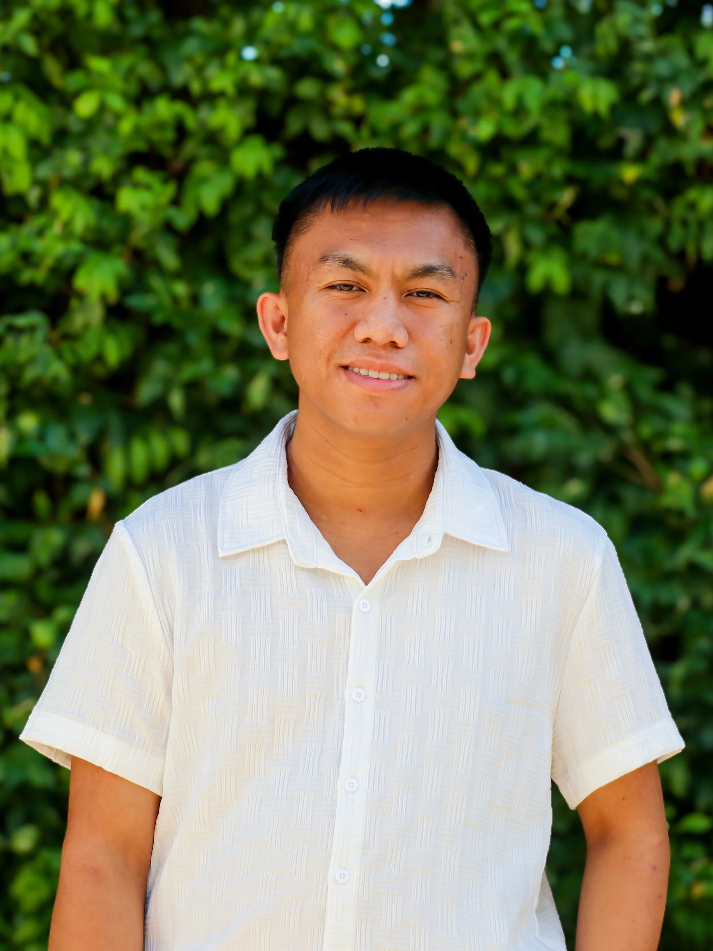 A young man with short black hair smiling outdoors, wearing a white button-up shirt, standing in front of green leafy bushes.