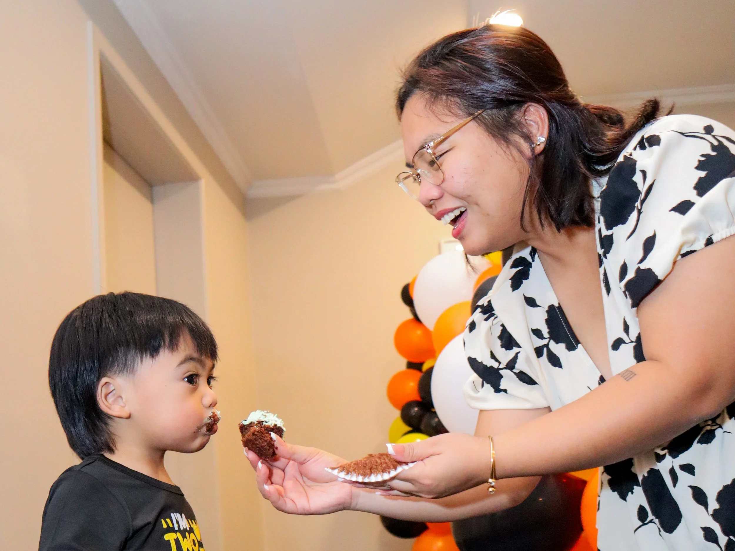 A woman wearing glasses and a cow-print dress is feeding a young boy a piece of chocolate cake at a birthday party with Halloween-themed balloon decorations in the background.