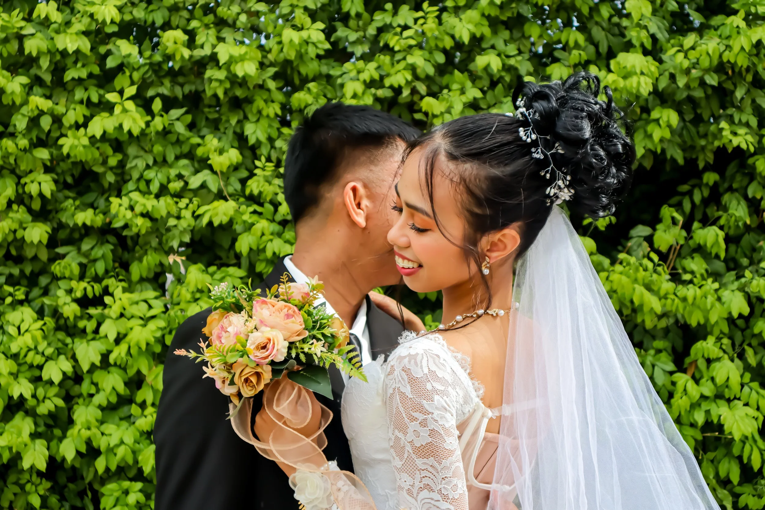 Bride and groom sharing a kiss outdoors, with green leafy background. The bride has styled hair with decorative hair accessories, wears a lace wedding dress, and holds a bouquet of pink and peach roses and greenery. The groom wears a black suit.