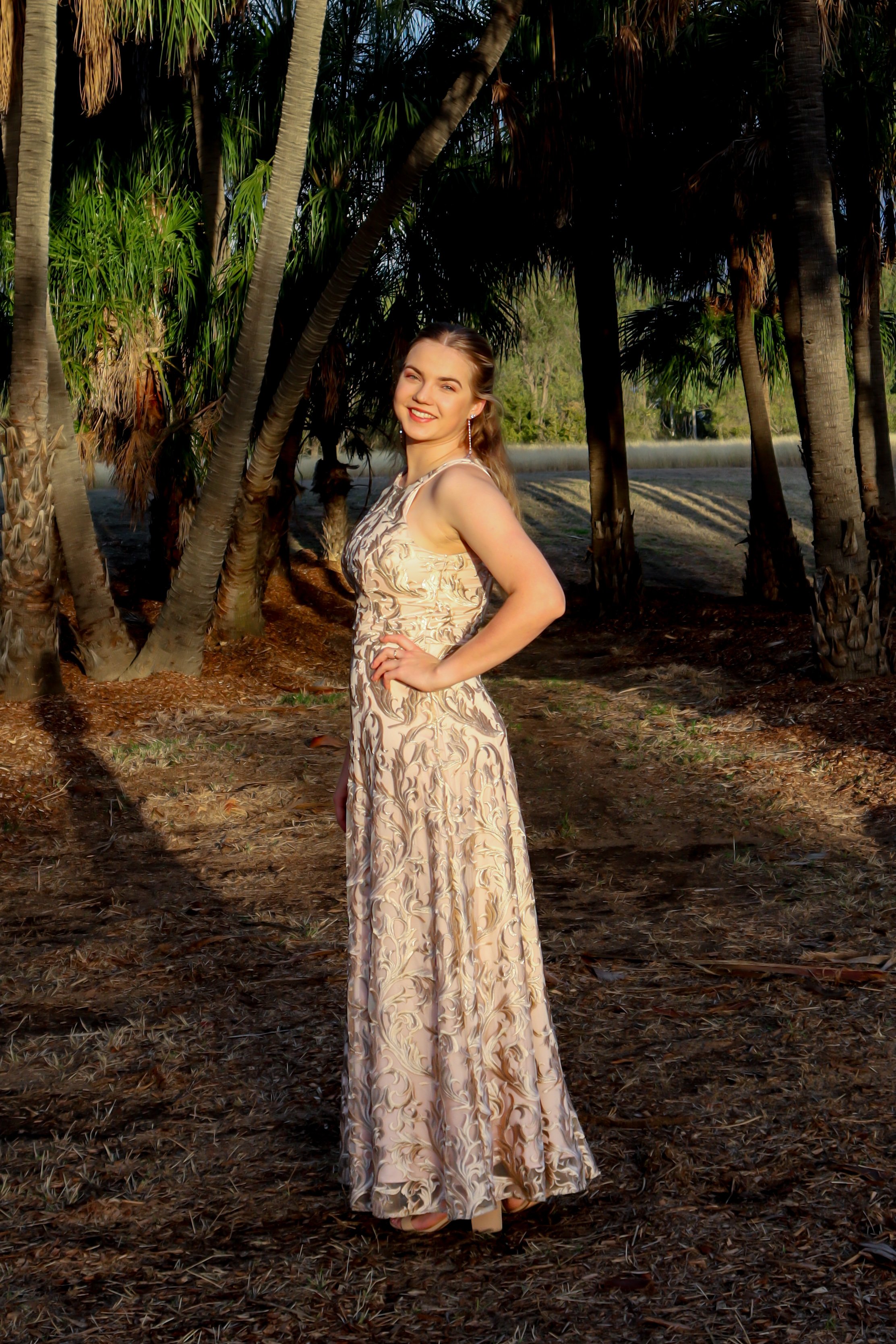 A woman in a long, patterned evening gown standing outdoors among palm trees, smiling with one hand on her hip.