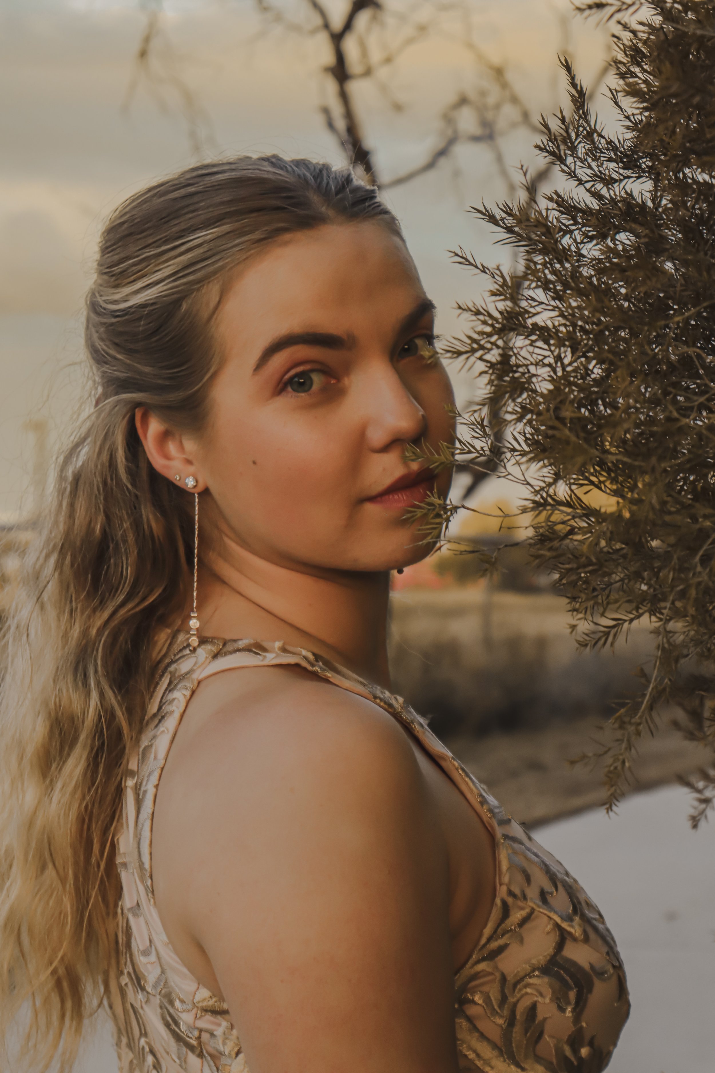 A young woman with long wavy blonde hair standing outdoors near a tree, looking over her shoulder at the camera during sunset.