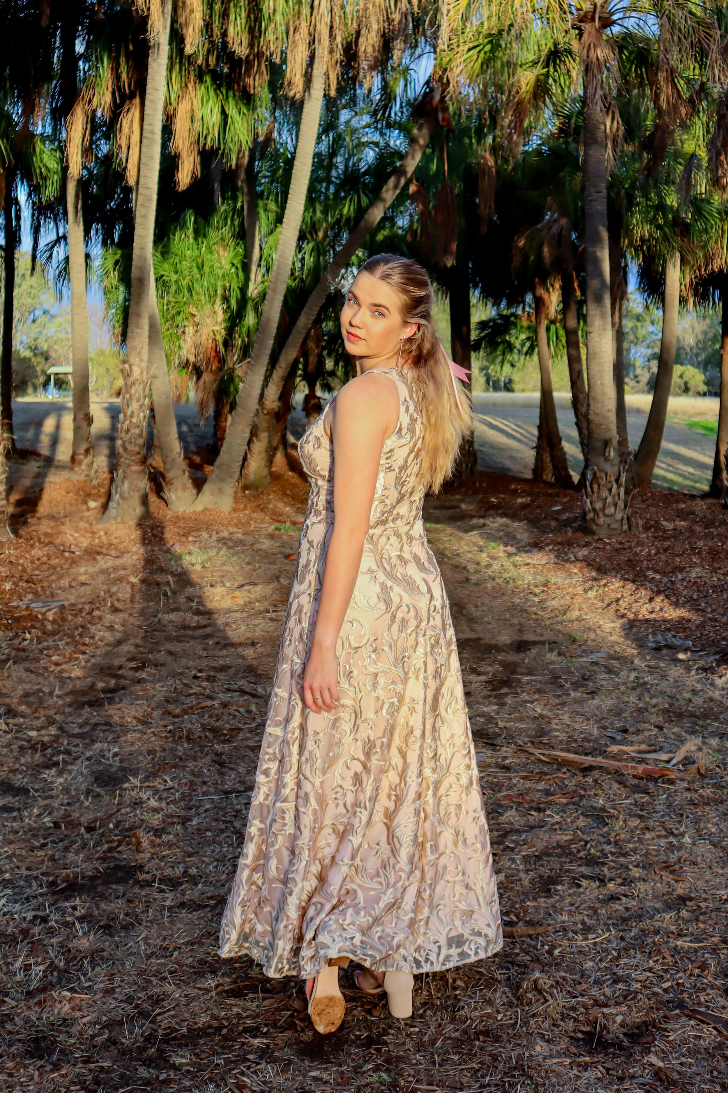 A woman in a sleeveless, long patterned dress standing outdoors among tall palm trees during sunset.