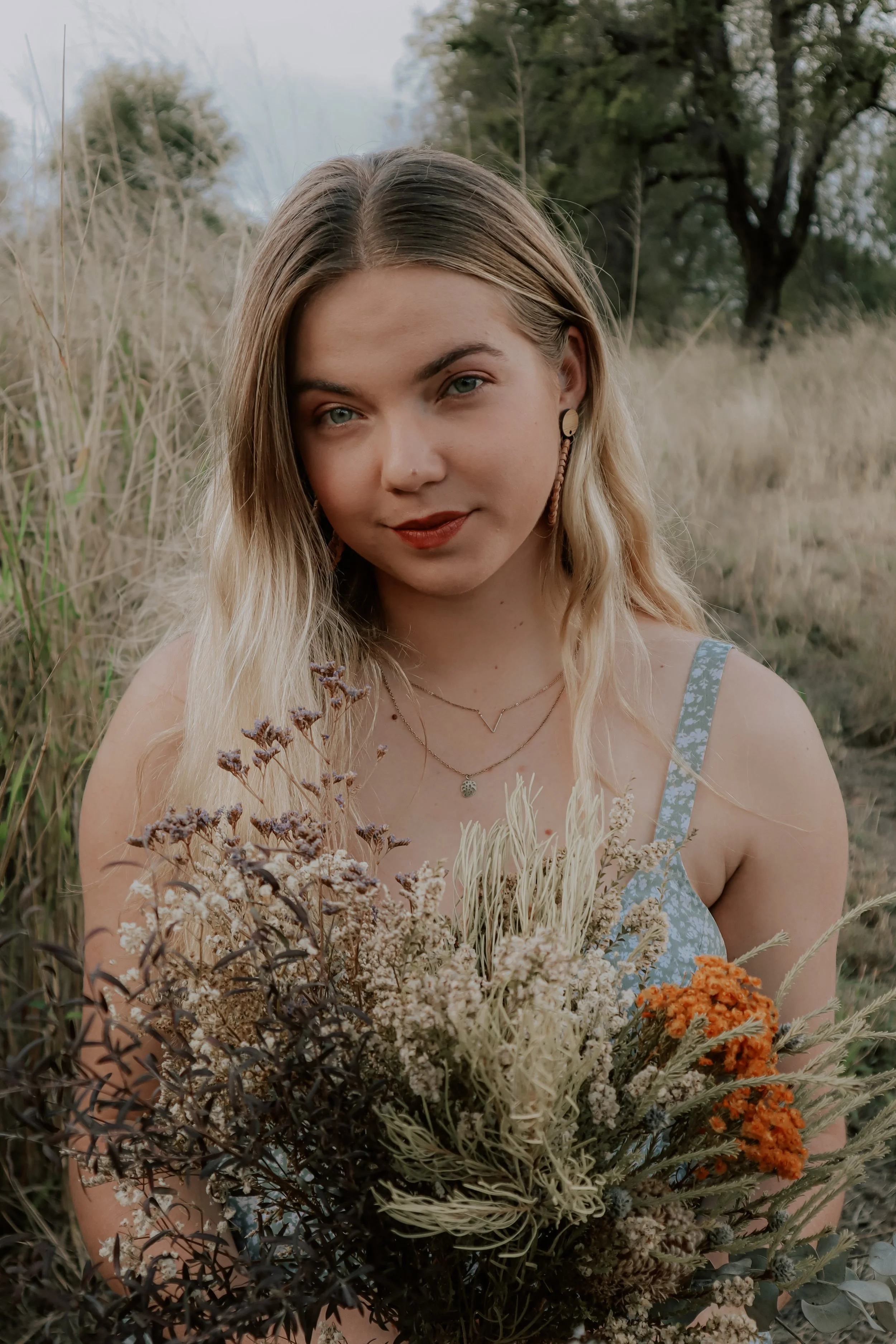 A young woman with blonde hair, wearing a blue floral sleeveless dress, holding a bouquet of dried wildflowers, standing outdoors among tall grasses with trees in the background.